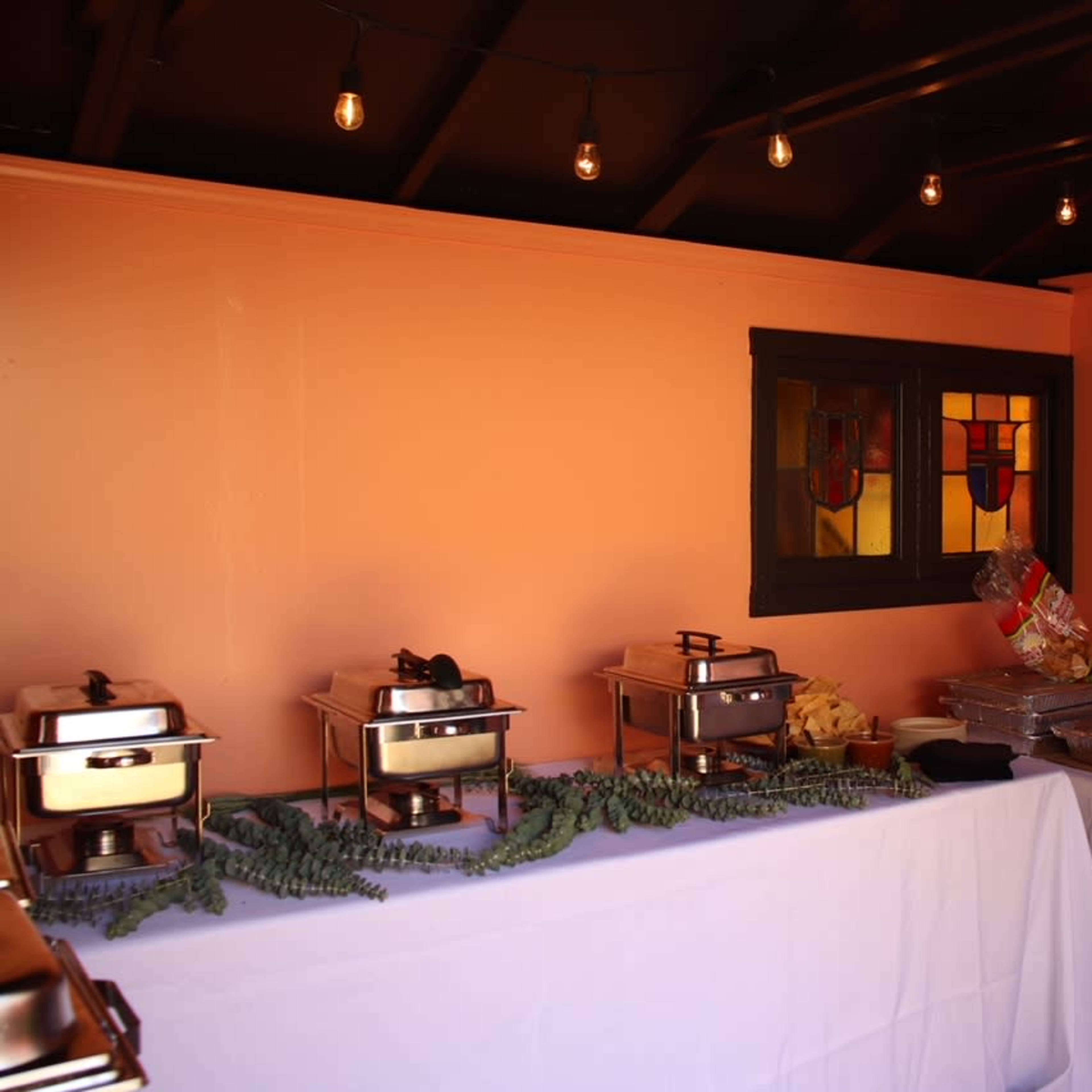 A buffet table is set up along a wall with chafing dishes, condiments, and a decorative arrangement of greenery.