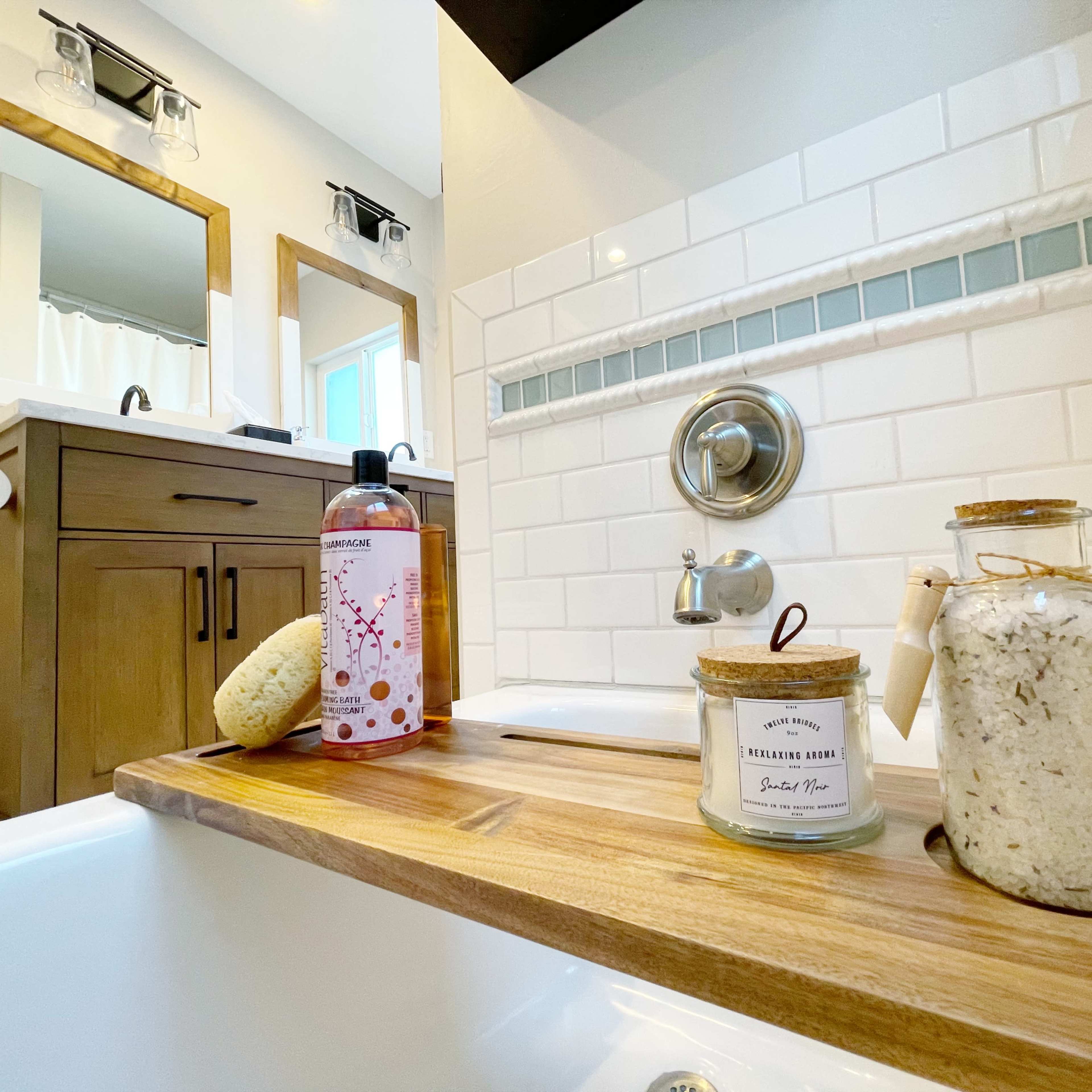 A well-lit bathroom with a wooden bath tray holding a sponge, a shampoo bottle, and a glass jar filled with bath salts, beside a white tiled bathtub.