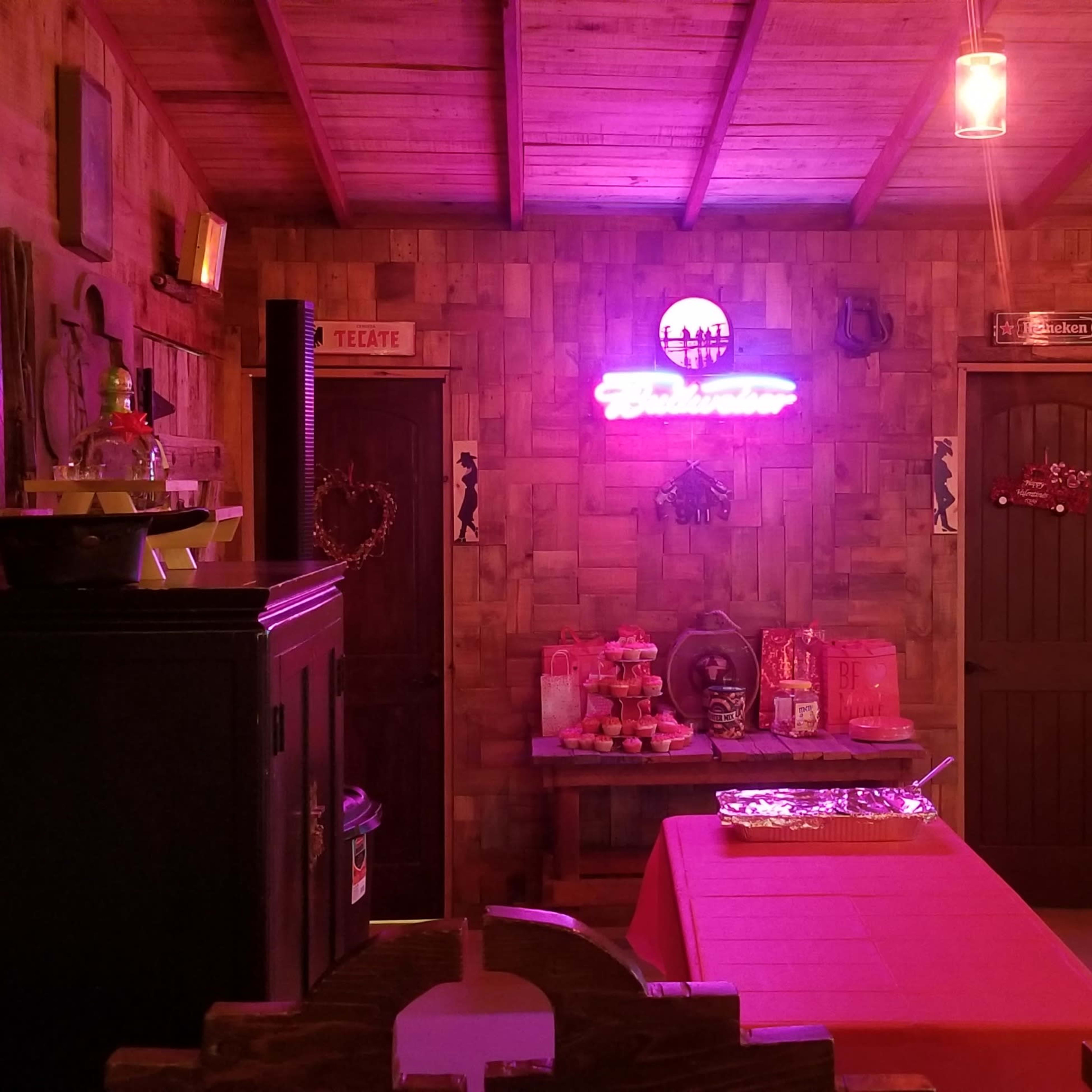The interior of a rustic bar with wooden walls, a neon sign, a television, and tables arranged for seating.