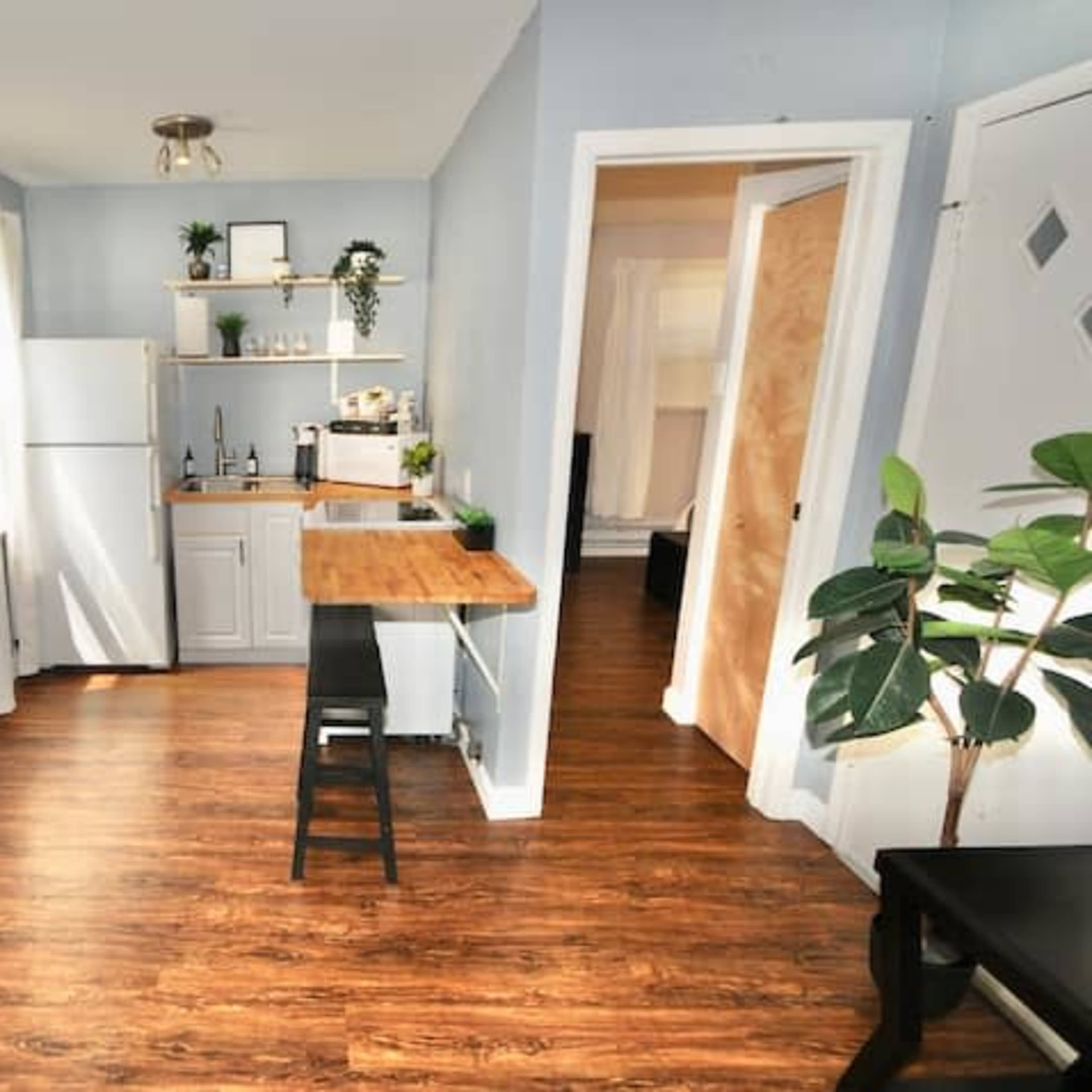 A small kitchen area with a refrigerator, a sink, a wooden table, and a doorway leading to another room.