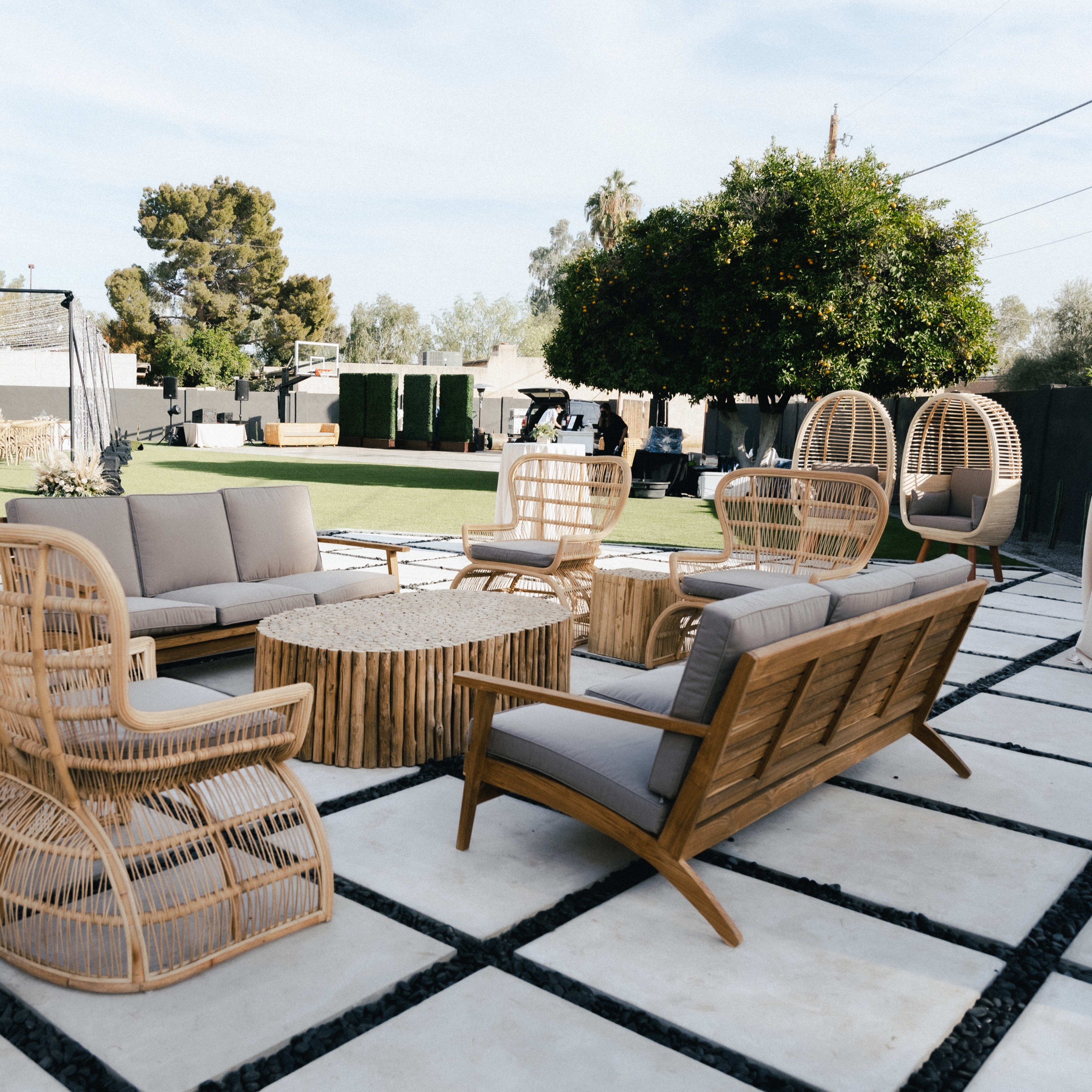 A modern outdoor lounge area featuring woven furniture with a central coffee table, surrounded by a grassy space and trees.
