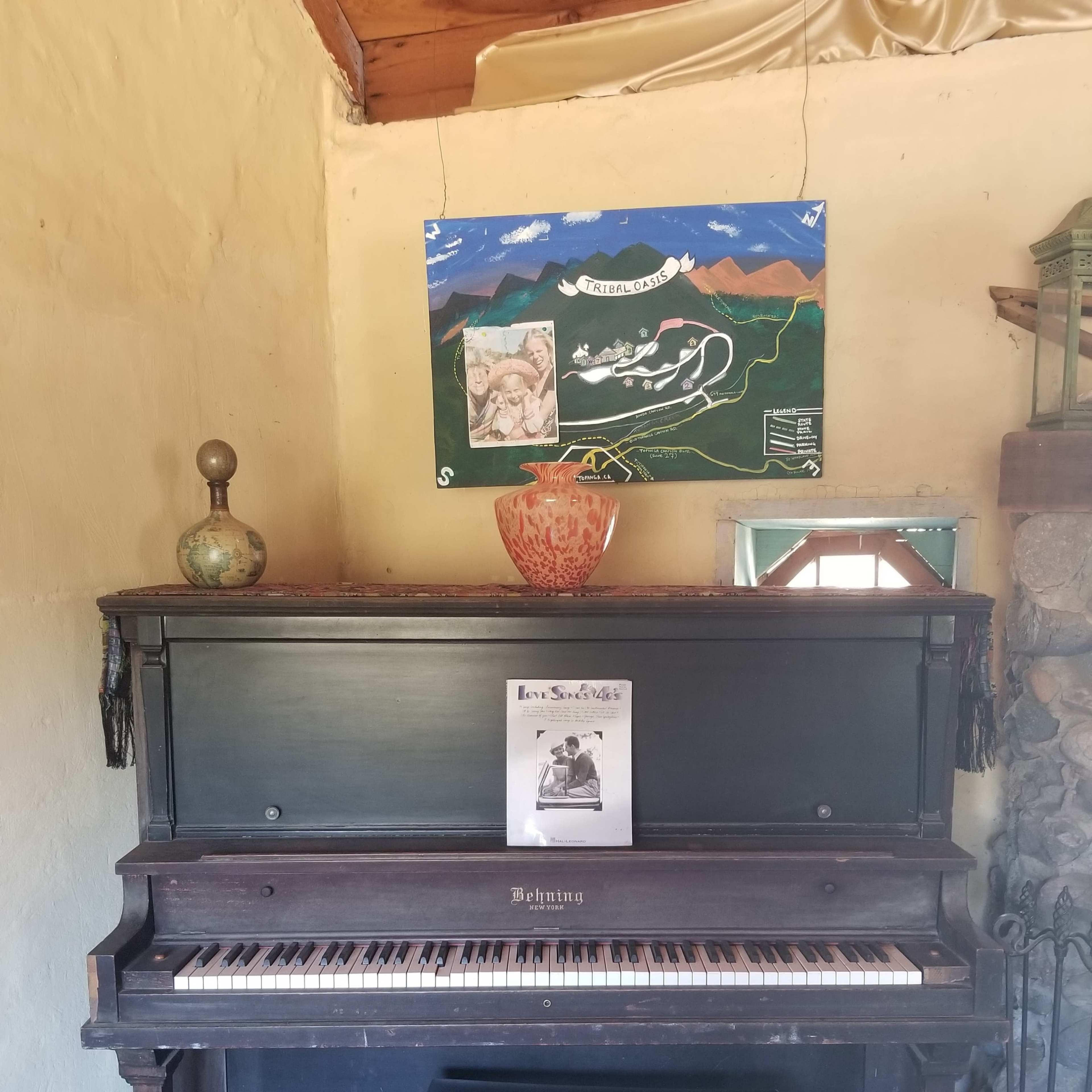 A vintage piano with a matching bench is positioned against a wall, accompanied by a decorative vase and a poster above featuring mountains and a map.