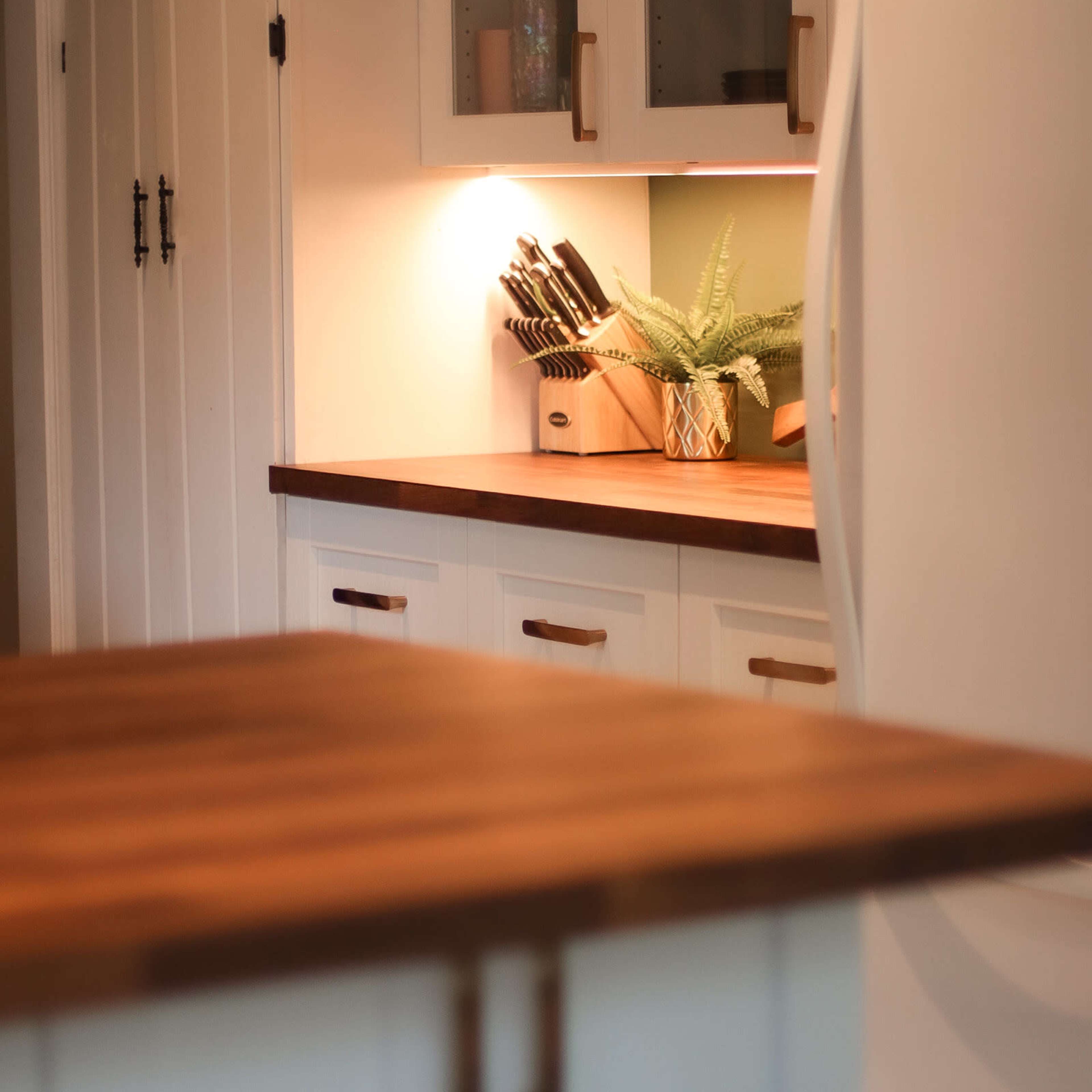 A clean kitchen features wooden countertops, white cabinets, and a well-organized knife rack illuminated by soft lighting.