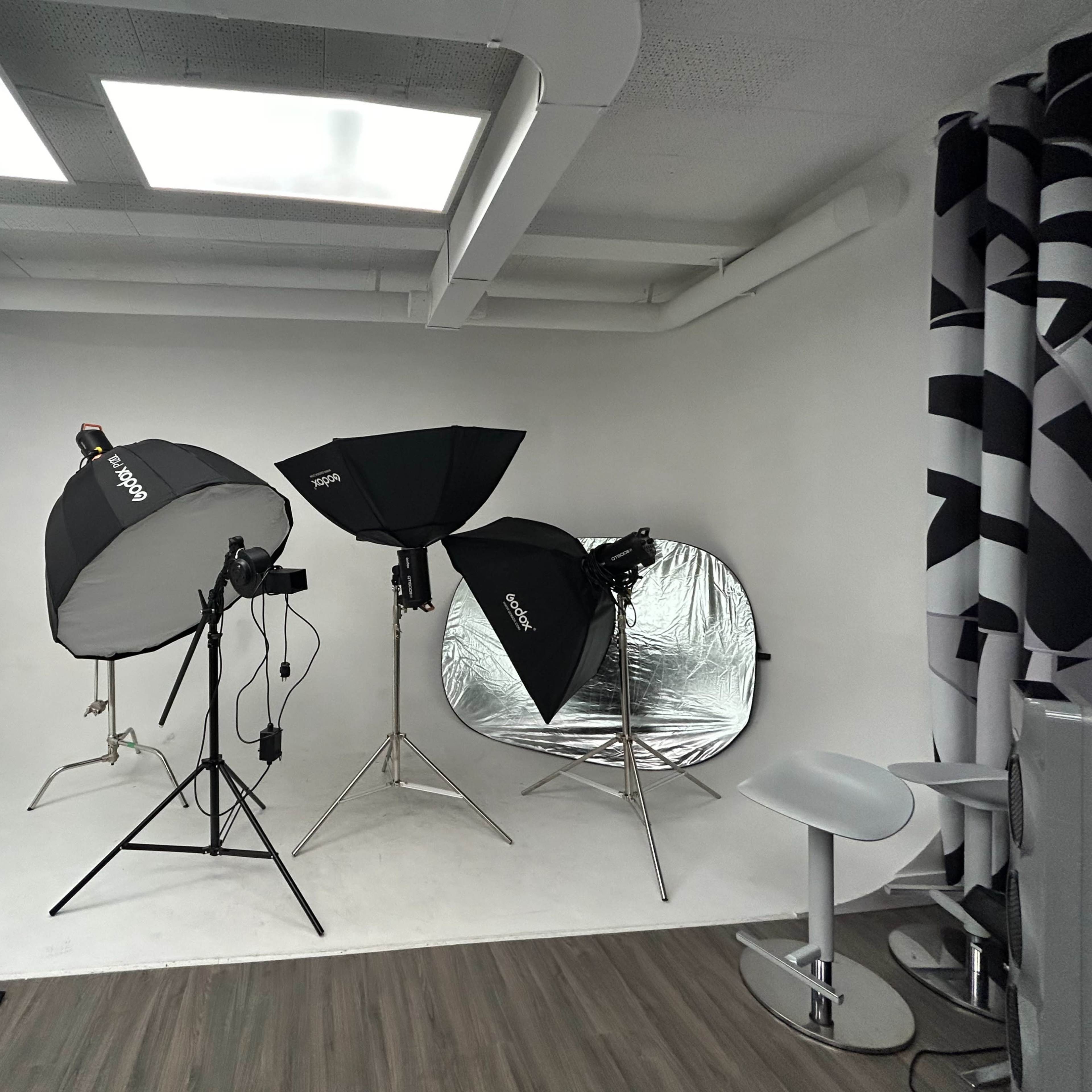 A photography studio with two softbox lights, a reflective umbrella, and a folding chair set against a white backdrop.