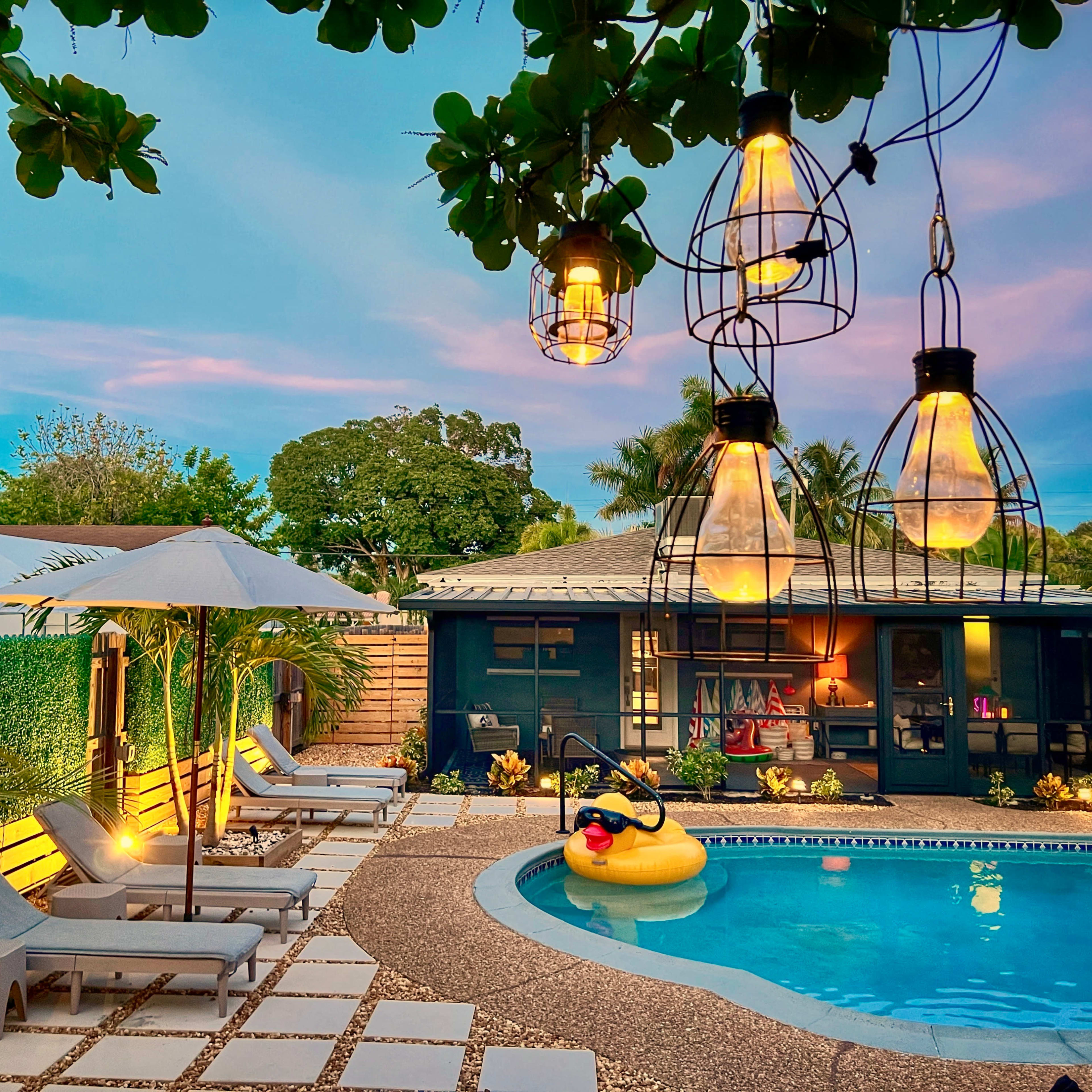 A well-lit backyard features a swimming pool surrounded by lounge chairs, a floating inflatable duck, and pendant lights hanging from overhead branches.