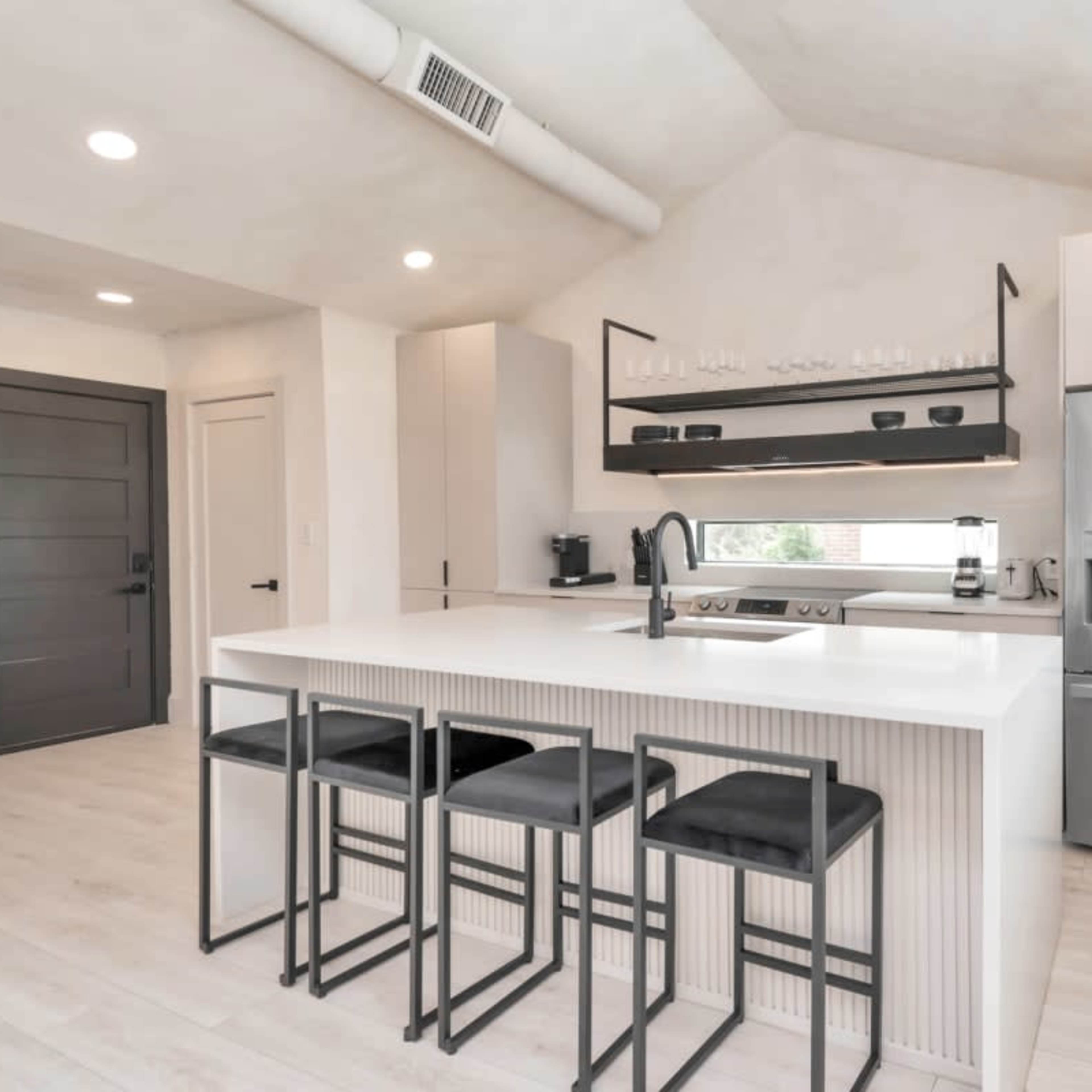 A modern kitchen with a white island, four black bar stools, stainless steel appliances, and ample natural light from a large window.