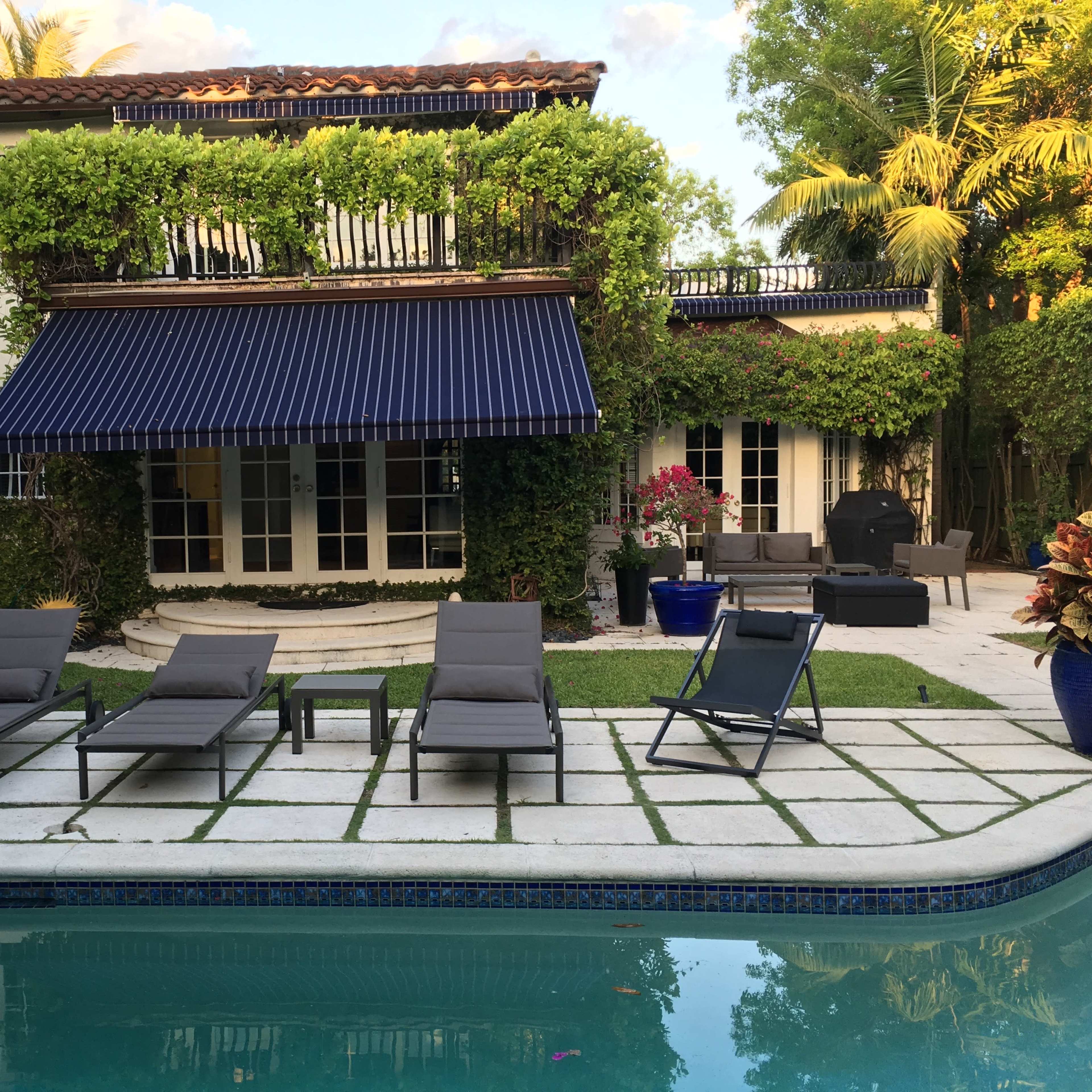 A lush backyard with a swimming pool, several lounge chairs arranged on a tiled patio, and a two-story house adorned with greenery and an awning.