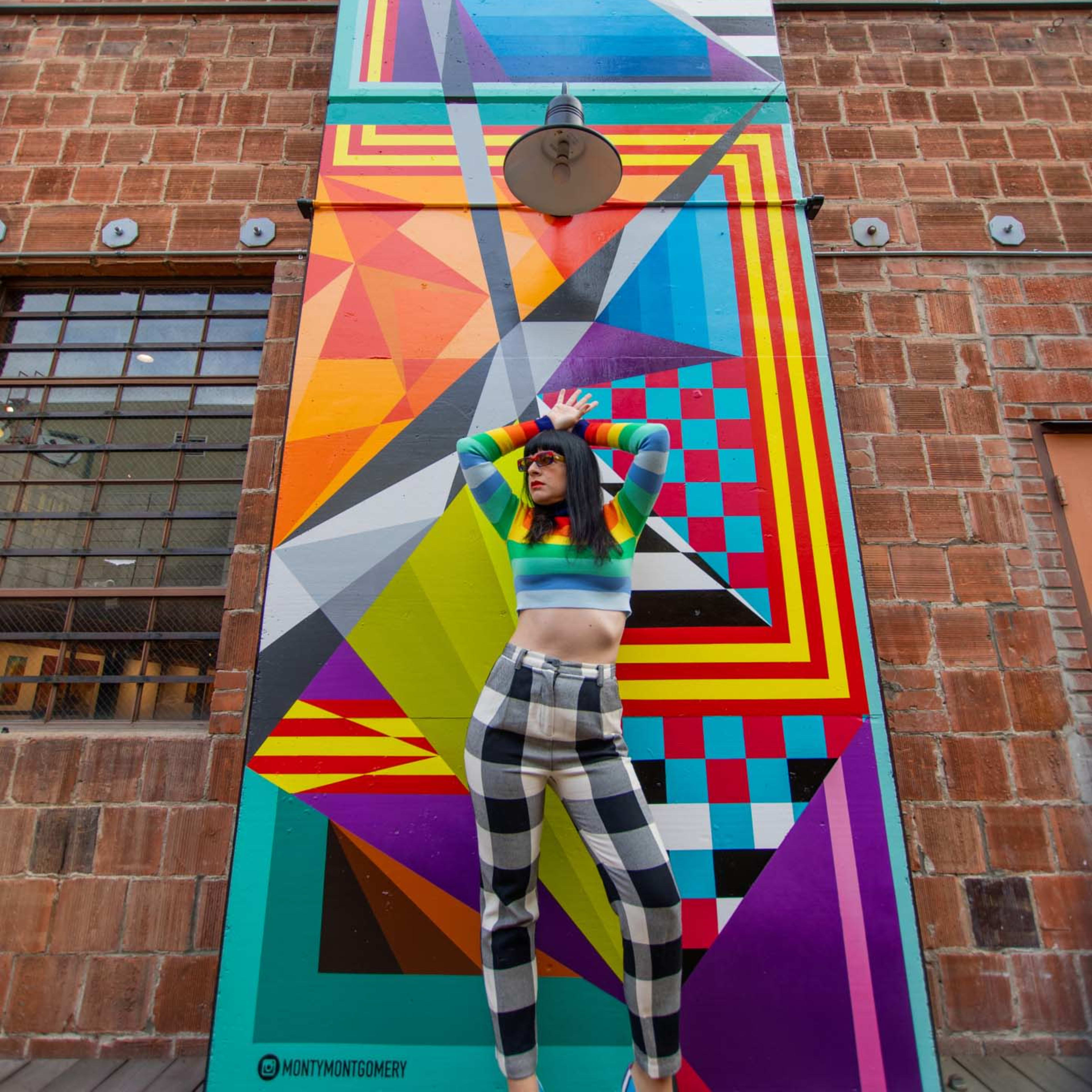 A woman poses in front of a colorful geometric mural, wearing a vibrant top and checkered pants.