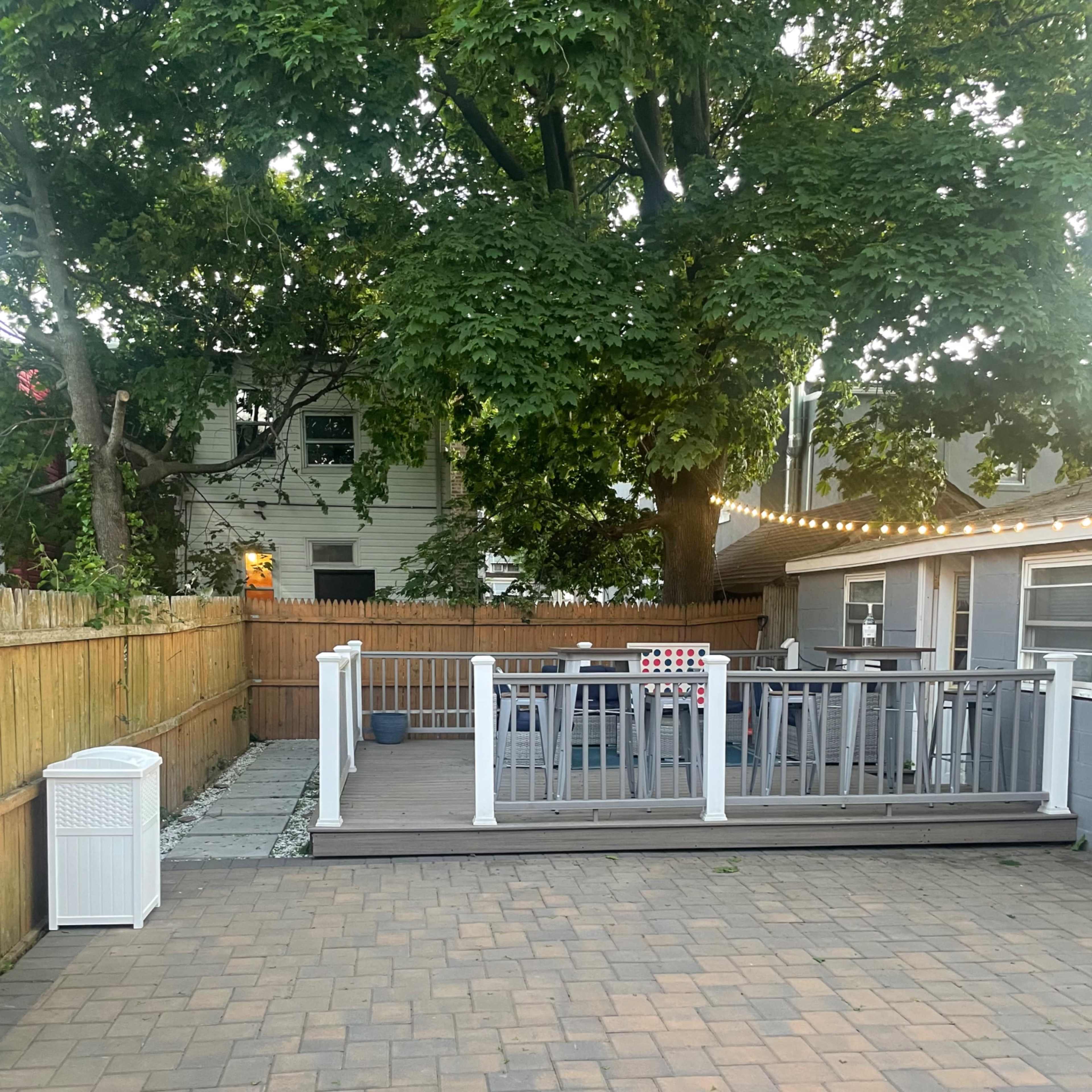 A patio area with a wooden deck featuring seating, surrounded by a fenced yard and trees.