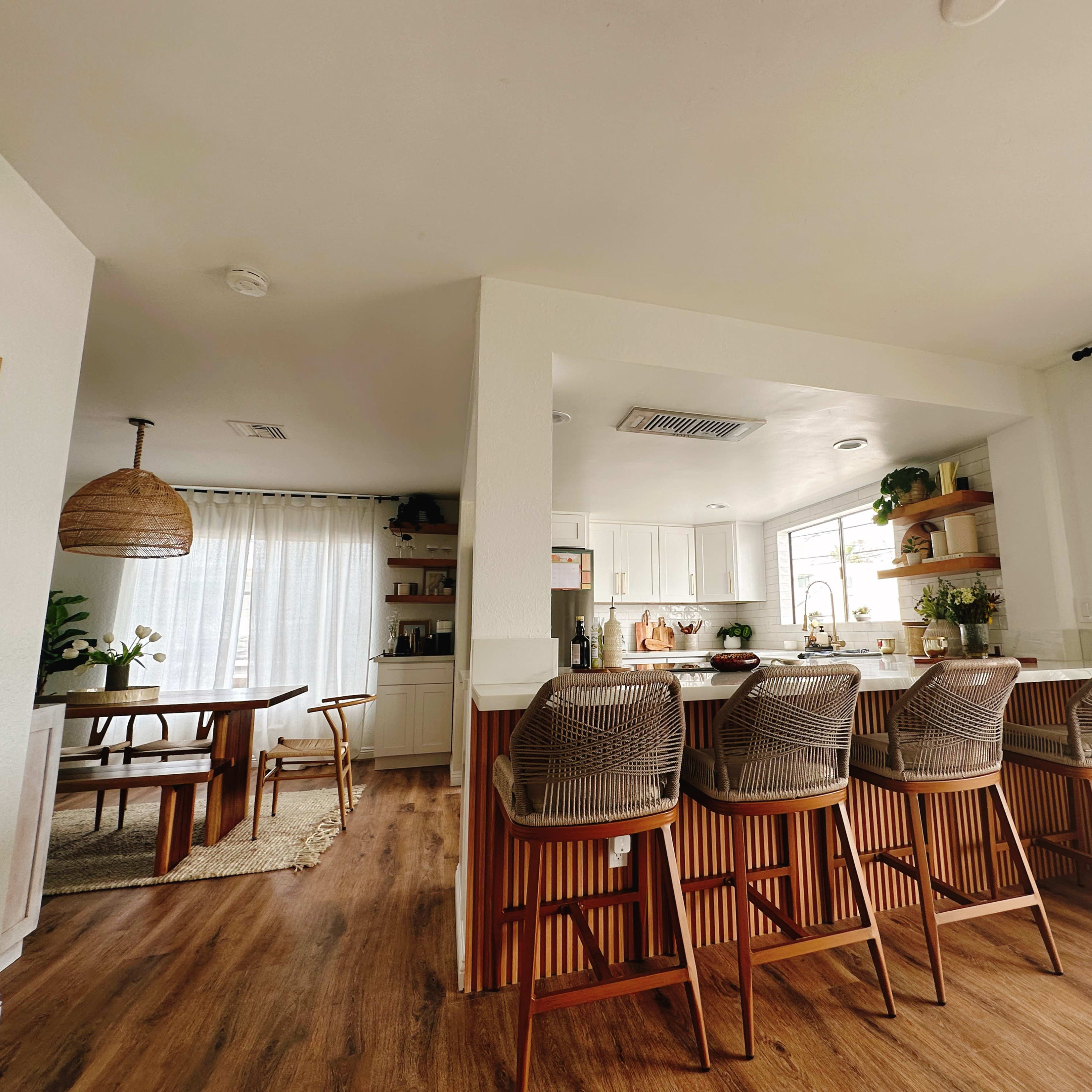 A modern kitchen with bar stools, a dining area, and large windows allowing natural light to enter.