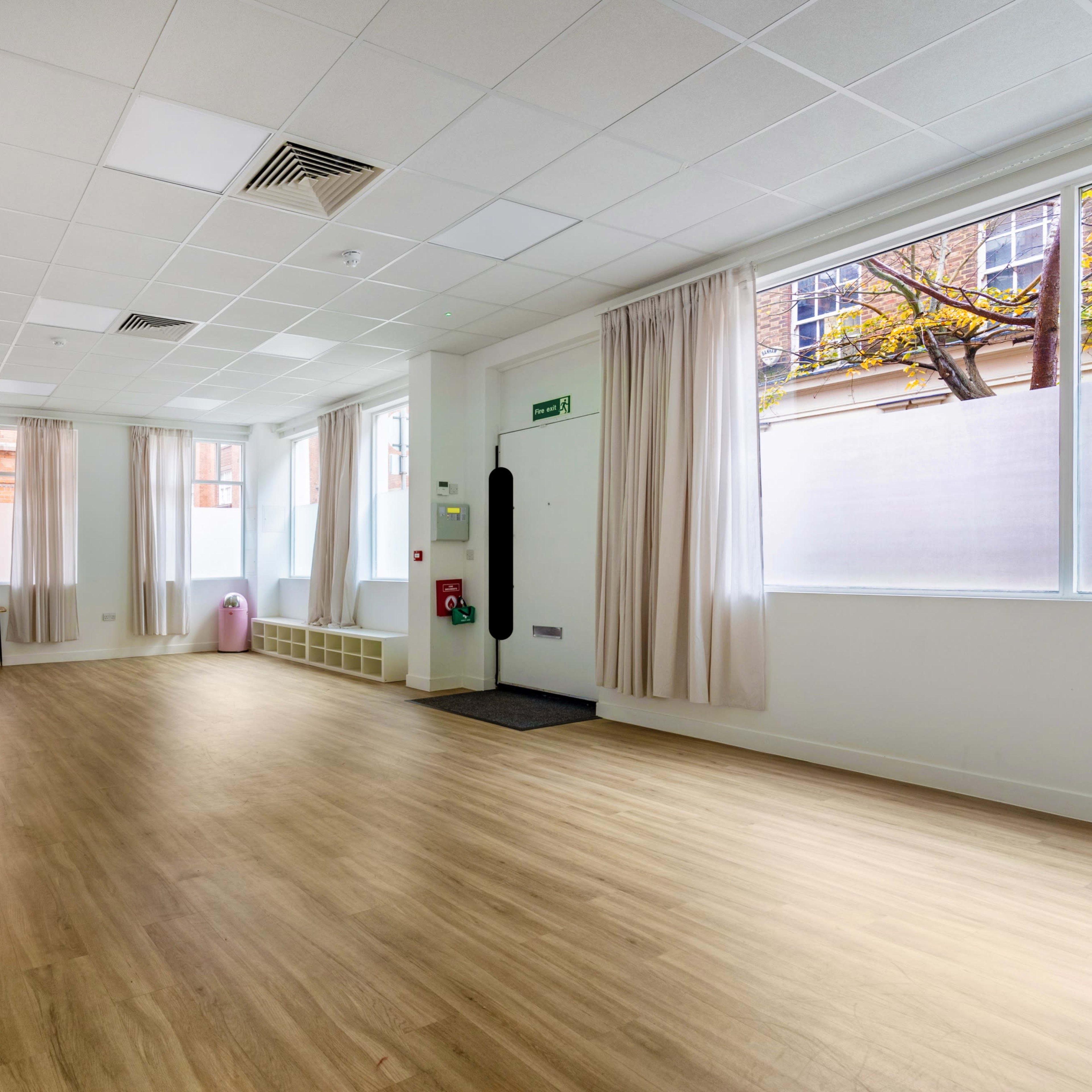 A spacious, well-lit room with large windows, wooden flooring, and a mirror along one wall.
