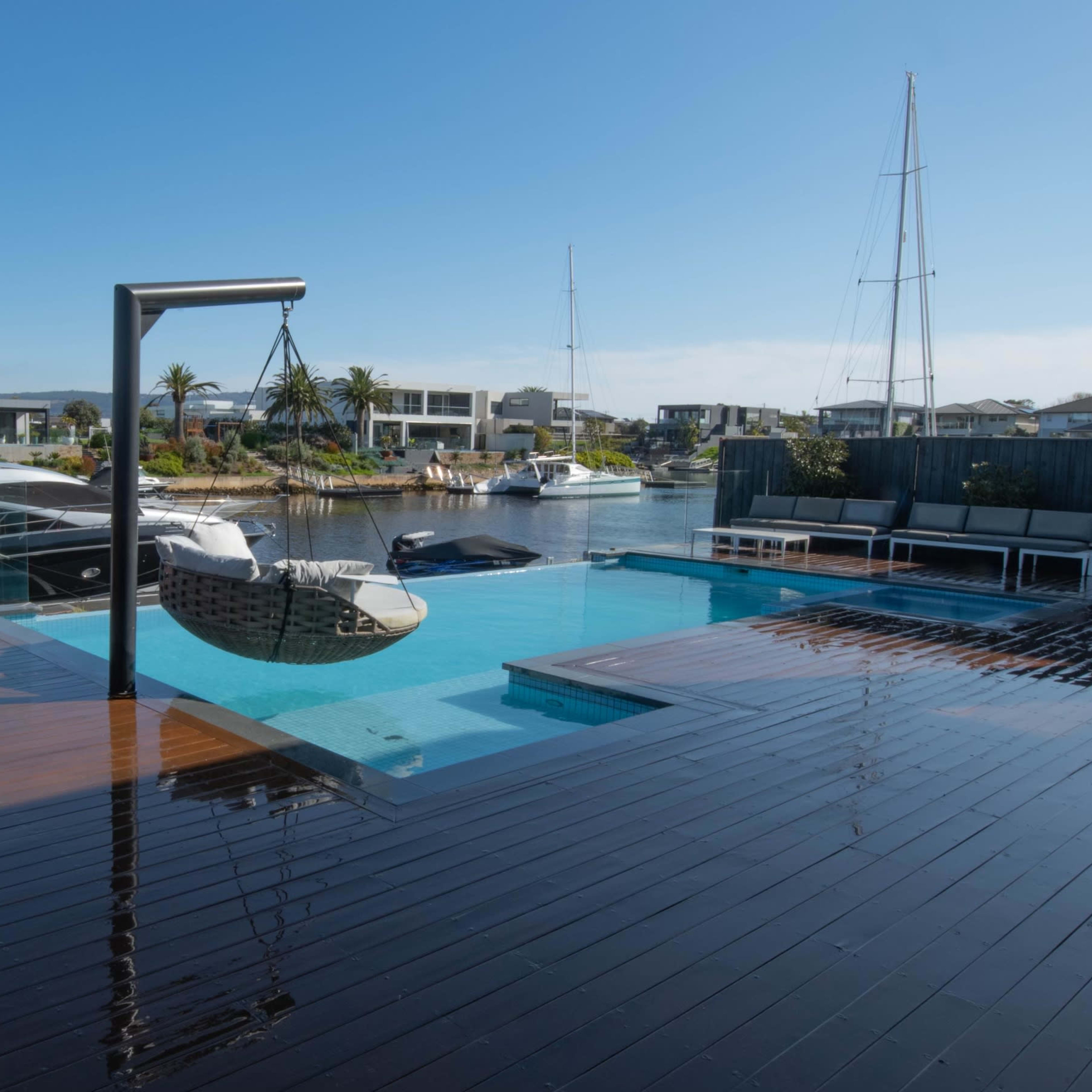 A modern waterfront property with a swimming pool, a hammock on a stand, and boats docked in a canal lined with houses.