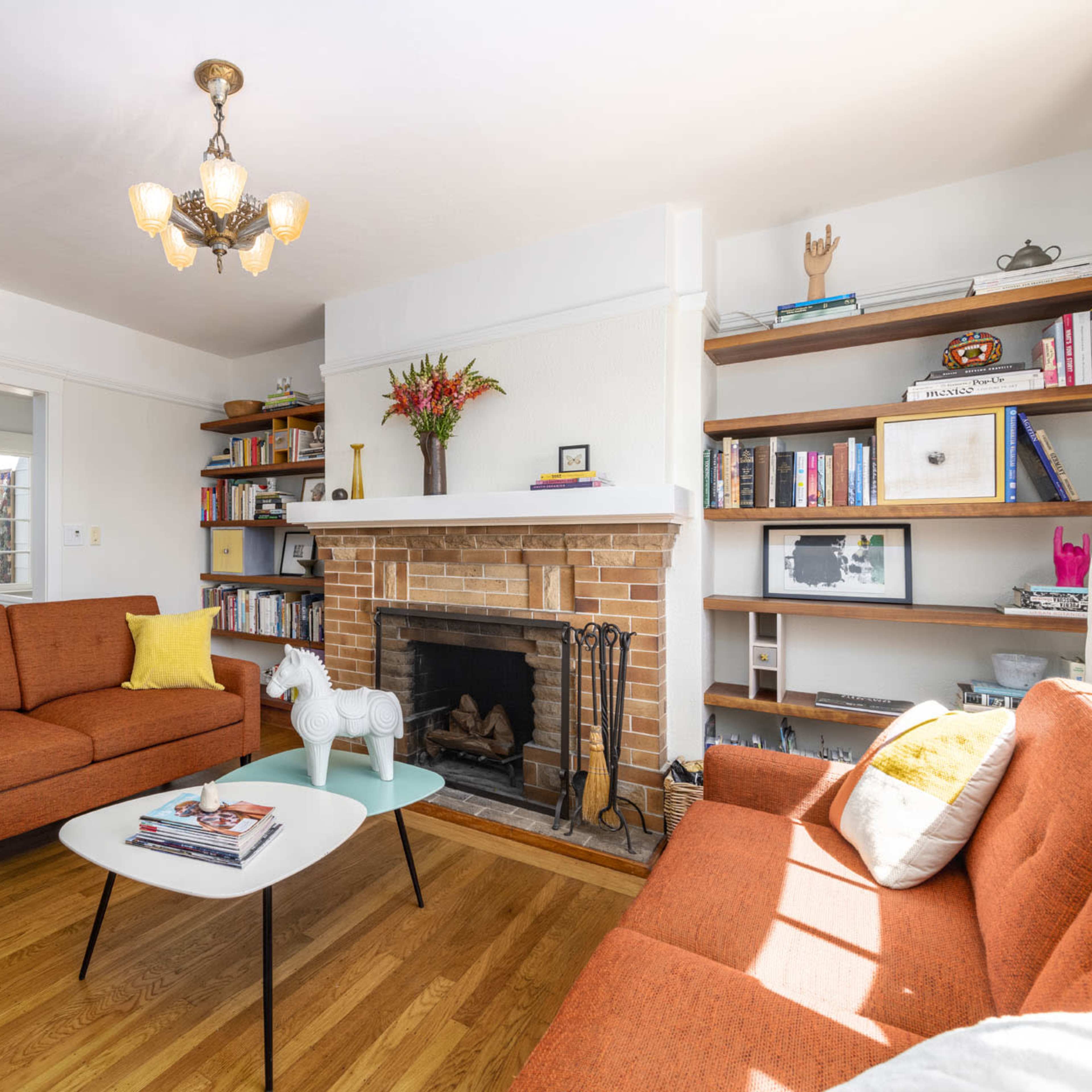 A living room with an orange upholstered sofa, a white coffee table, a brick fireplace, and shelves filled with books and decorative items.