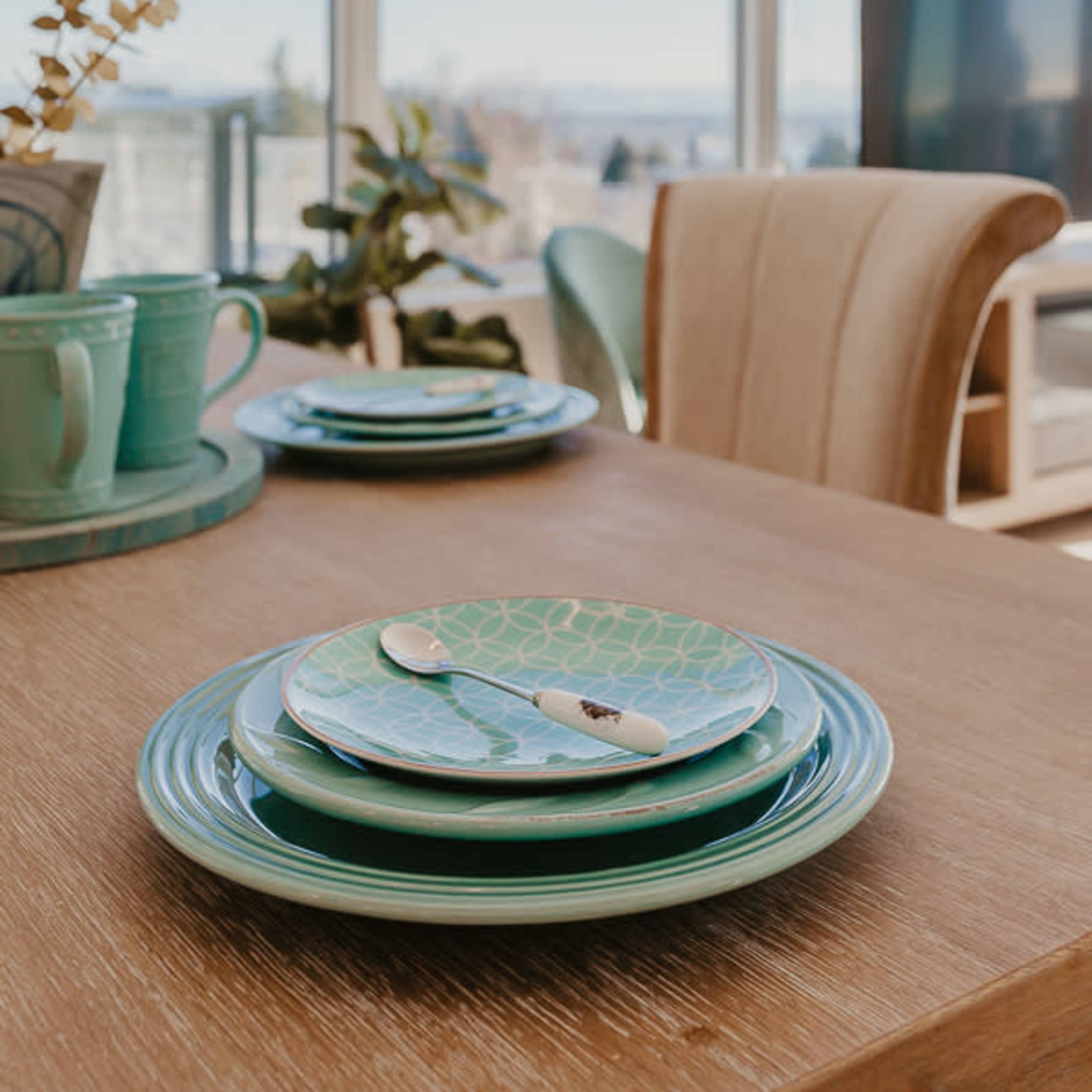 A dining table with stacked green plates and utensils is set against a backdrop of large windows offering a view of the outdoors.