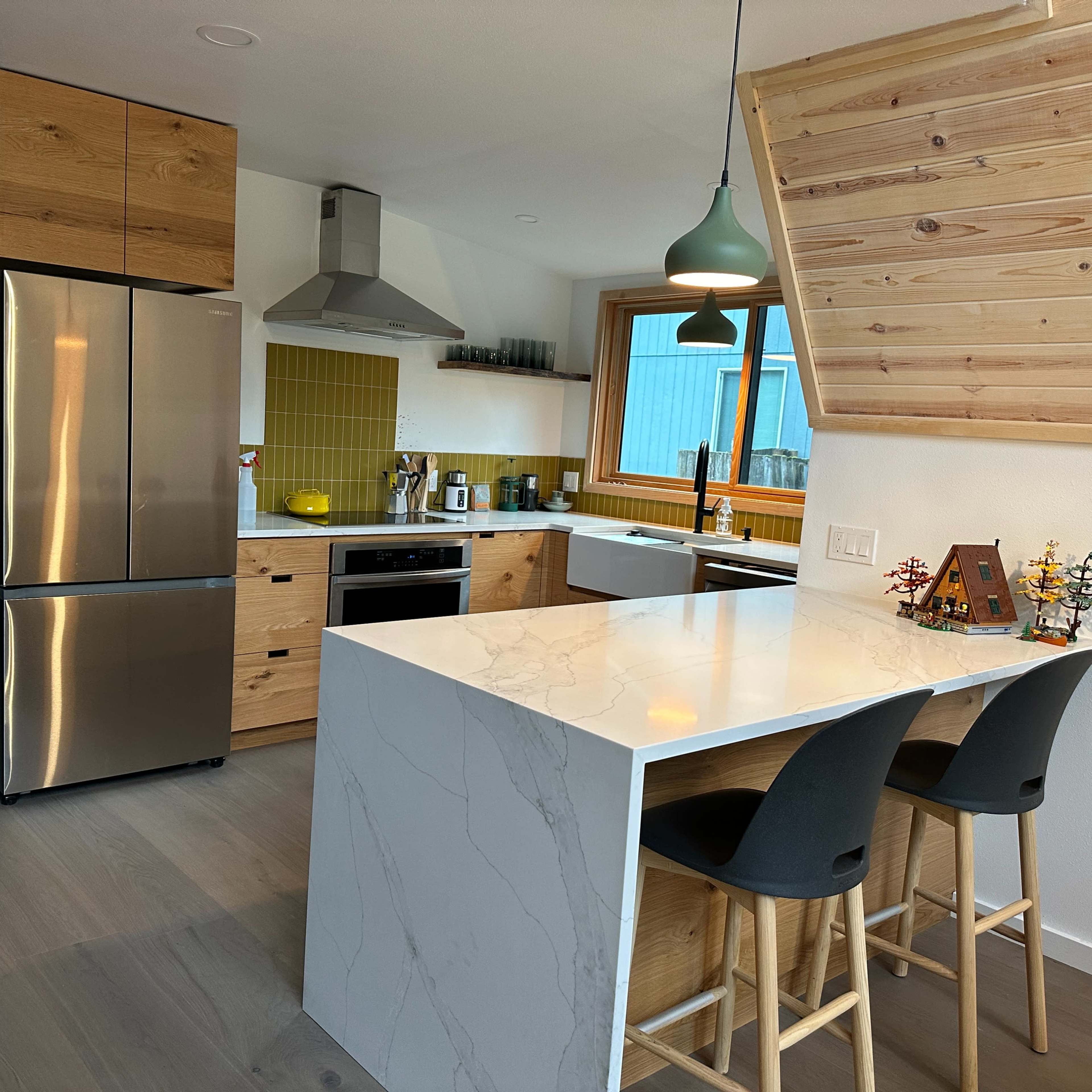 A modern kitchen featuring stainless steel appliances, a marble-topped island with seating, and wooden cabinetry against a bright yellow backsplash.