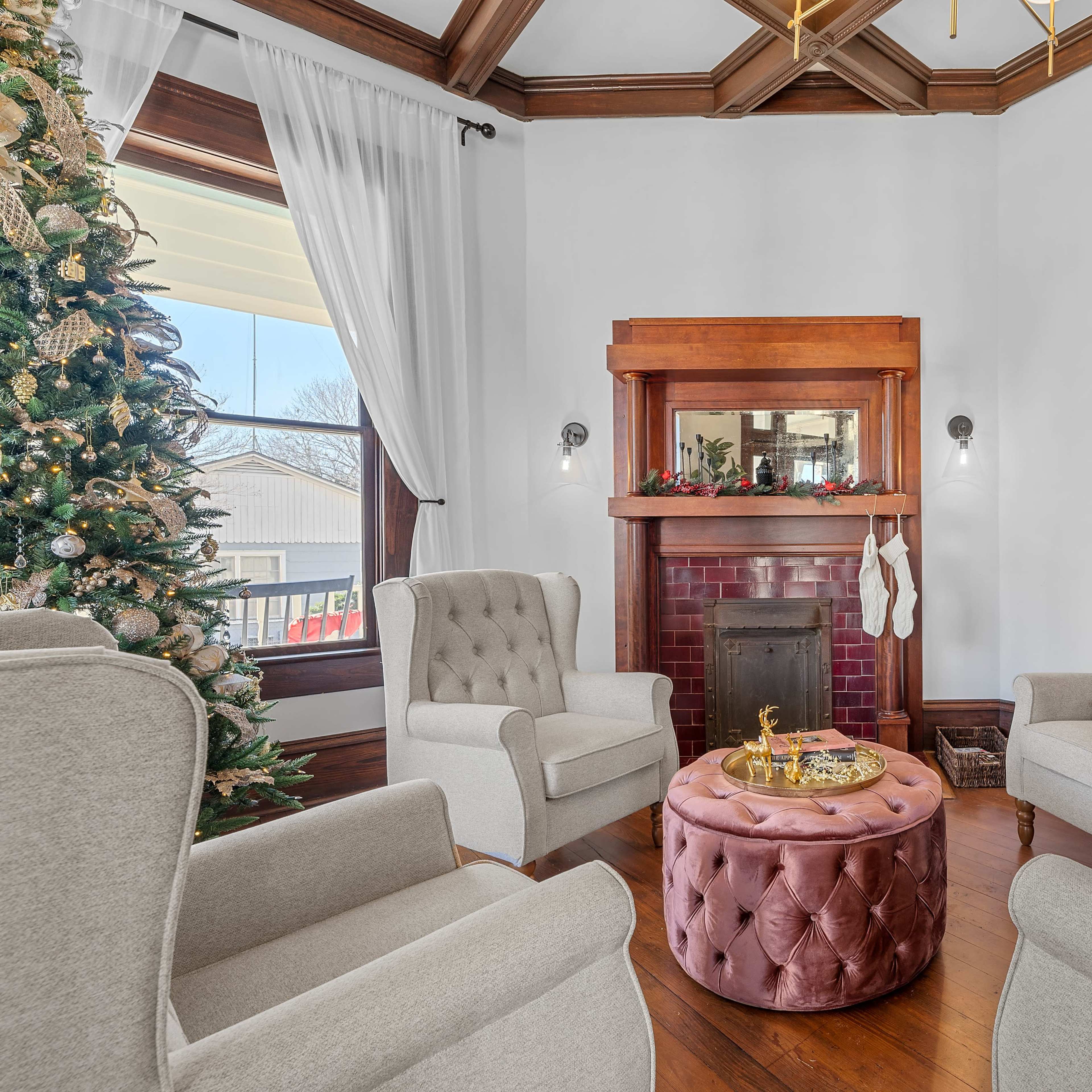 A well-furnished living room featuring a Christmas tree, a fireplace with decorative stockings, and four armchairs arranged around a round, tufted table.