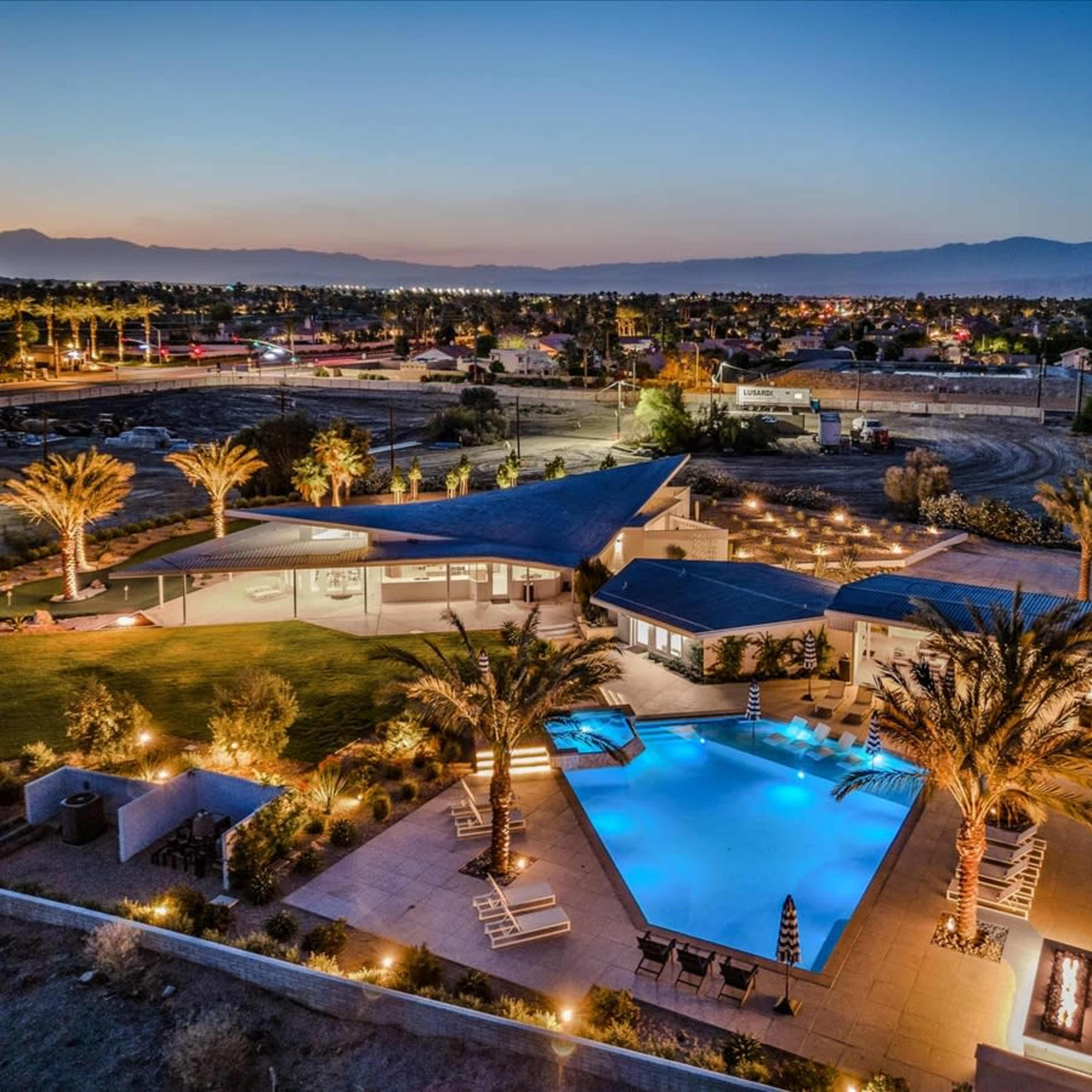 A modern home with a large swimming pool and palm trees is illuminated at dusk, surrounded by open land and distant mountains.