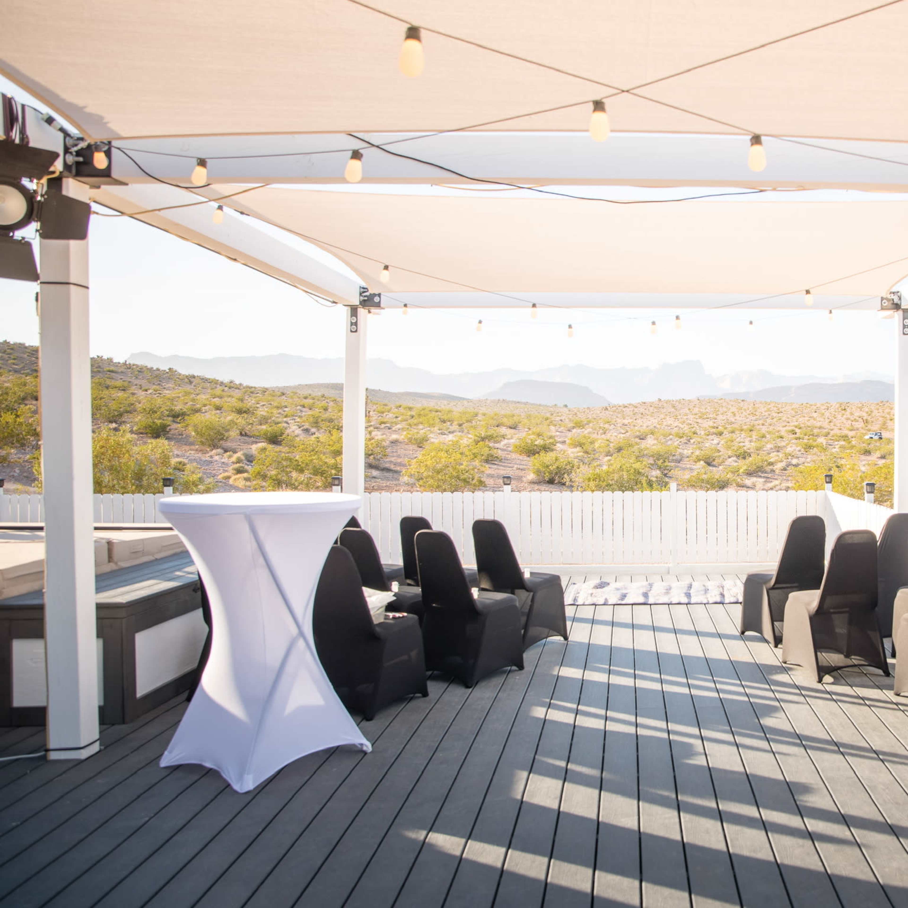 A terrace area features black chairs and white cocktail tables under a shaded canopy with string lights, overlooking a desert landscape.