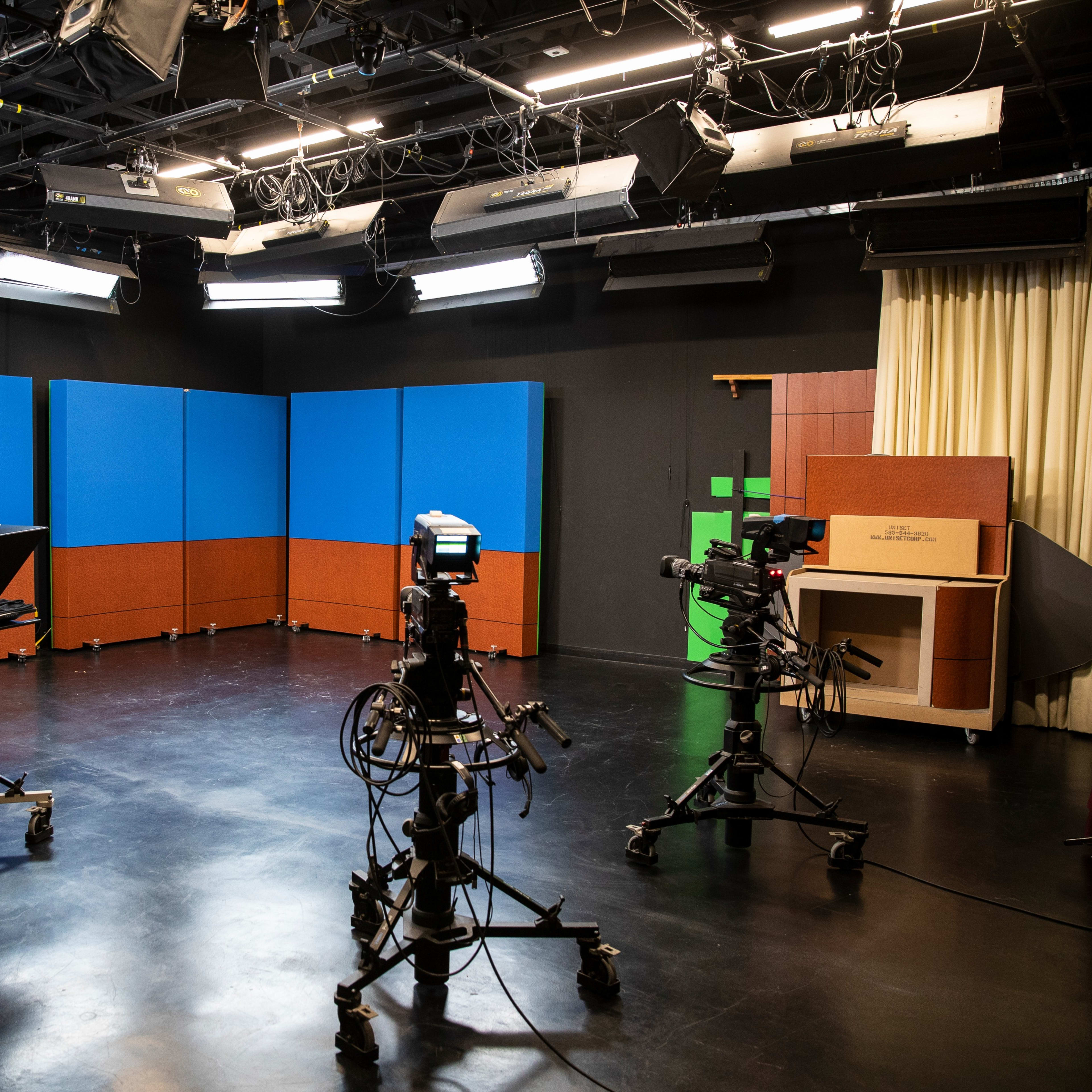 A television studio is set up with multiple cameras facing a blue backdrop and equipment, surrounded by studio lights and seating.