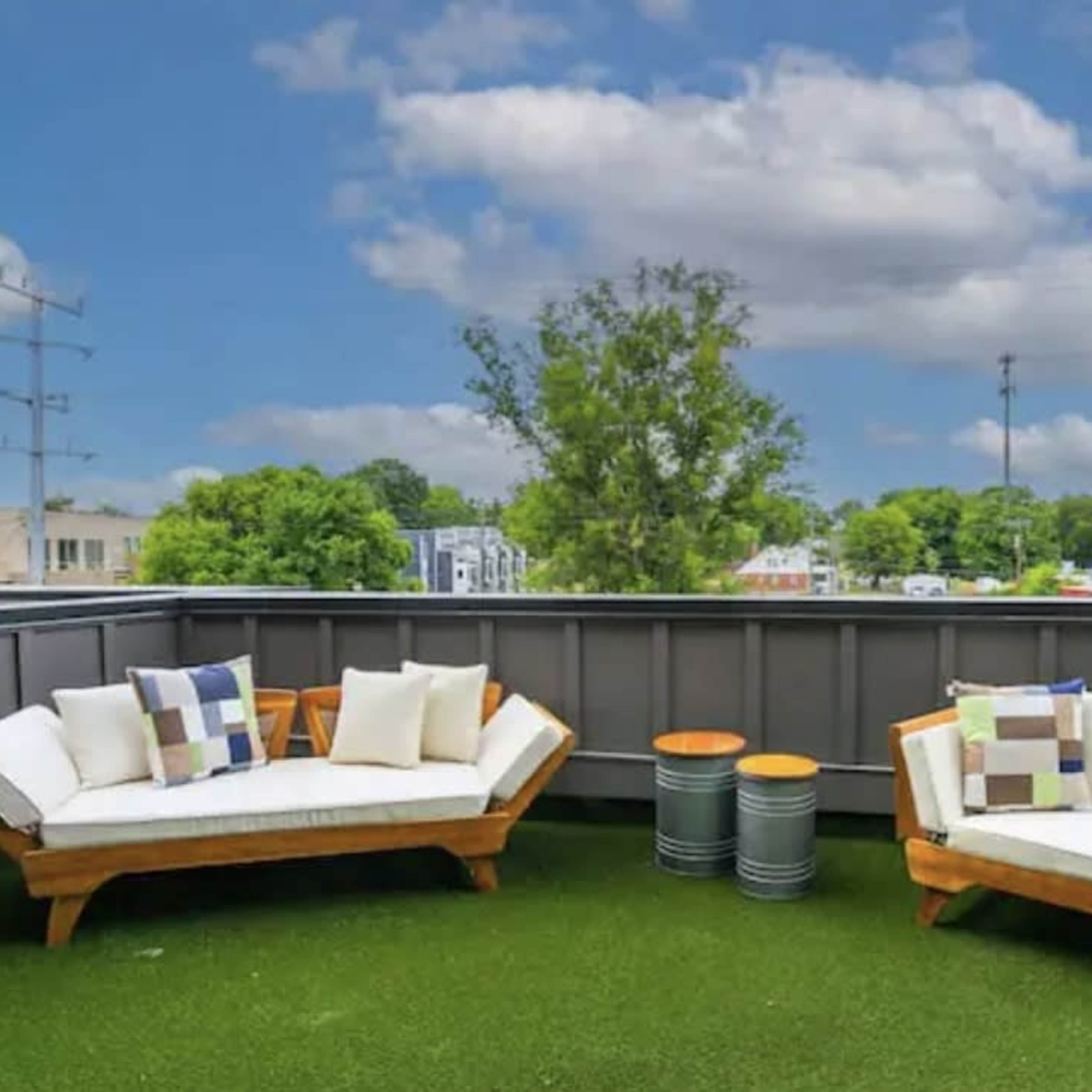 A balcony features two wooden sofas with white cushions, flanked by two small tables, all set against a backdrop of green grass and a blue sky with clouds.
