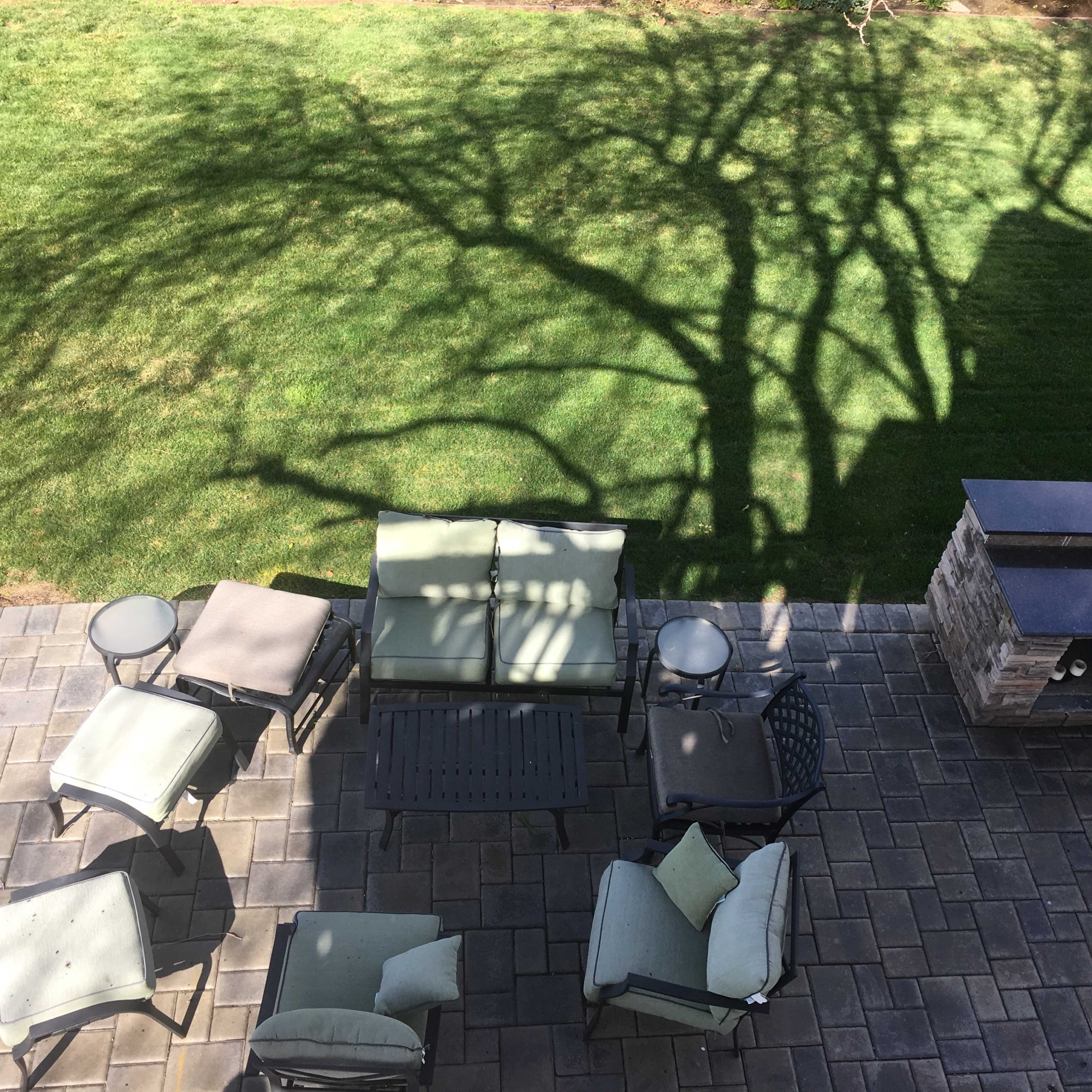 A patio area with several pieces of outdoor furniture, including a sofa, chairs, and a table, cast in the shadows of a nearby tree in a grassy yard.
