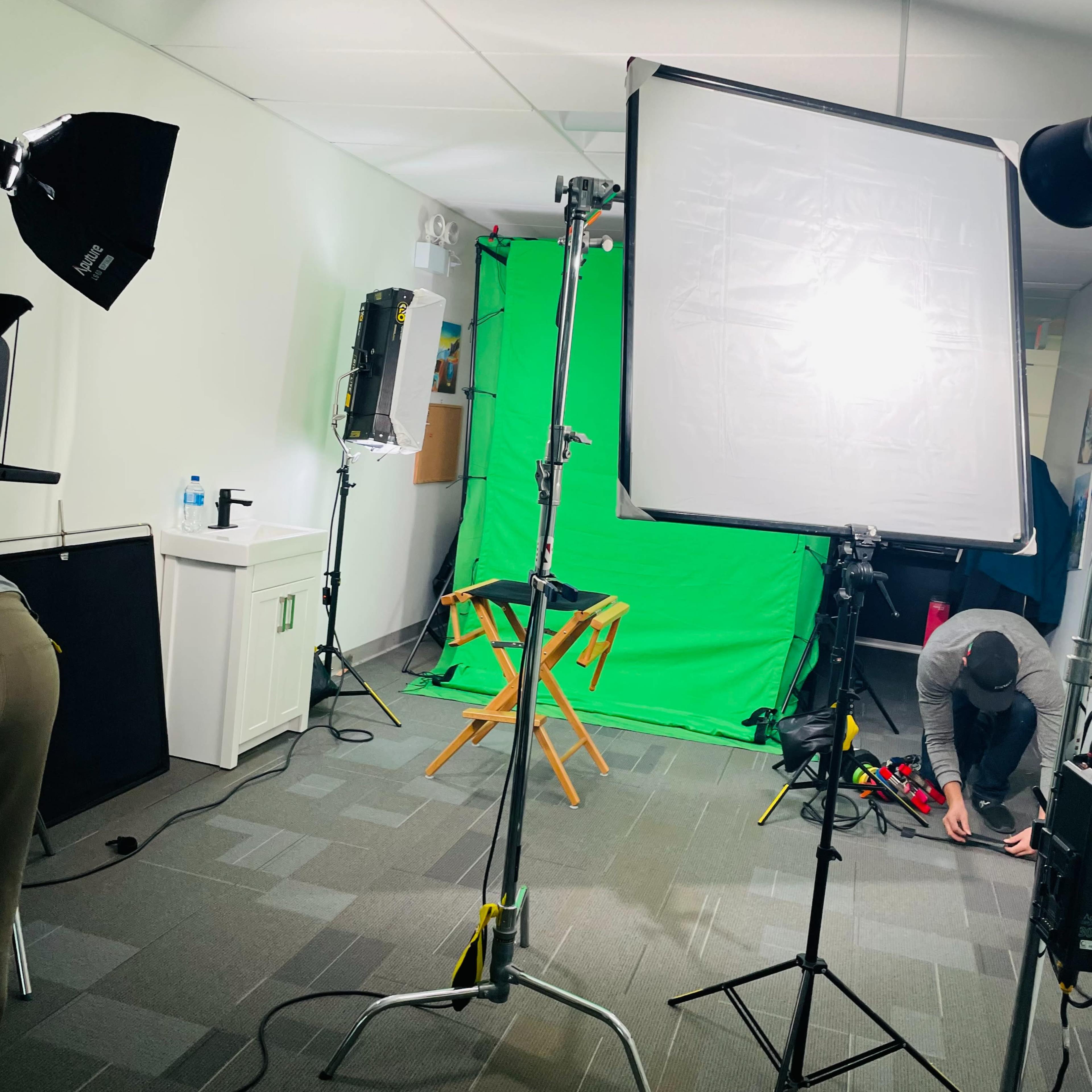 A photo studio setup featuring a green screen backdrop, multiple light sources, and a director's chair, with a person adjusting equipment on the floor.