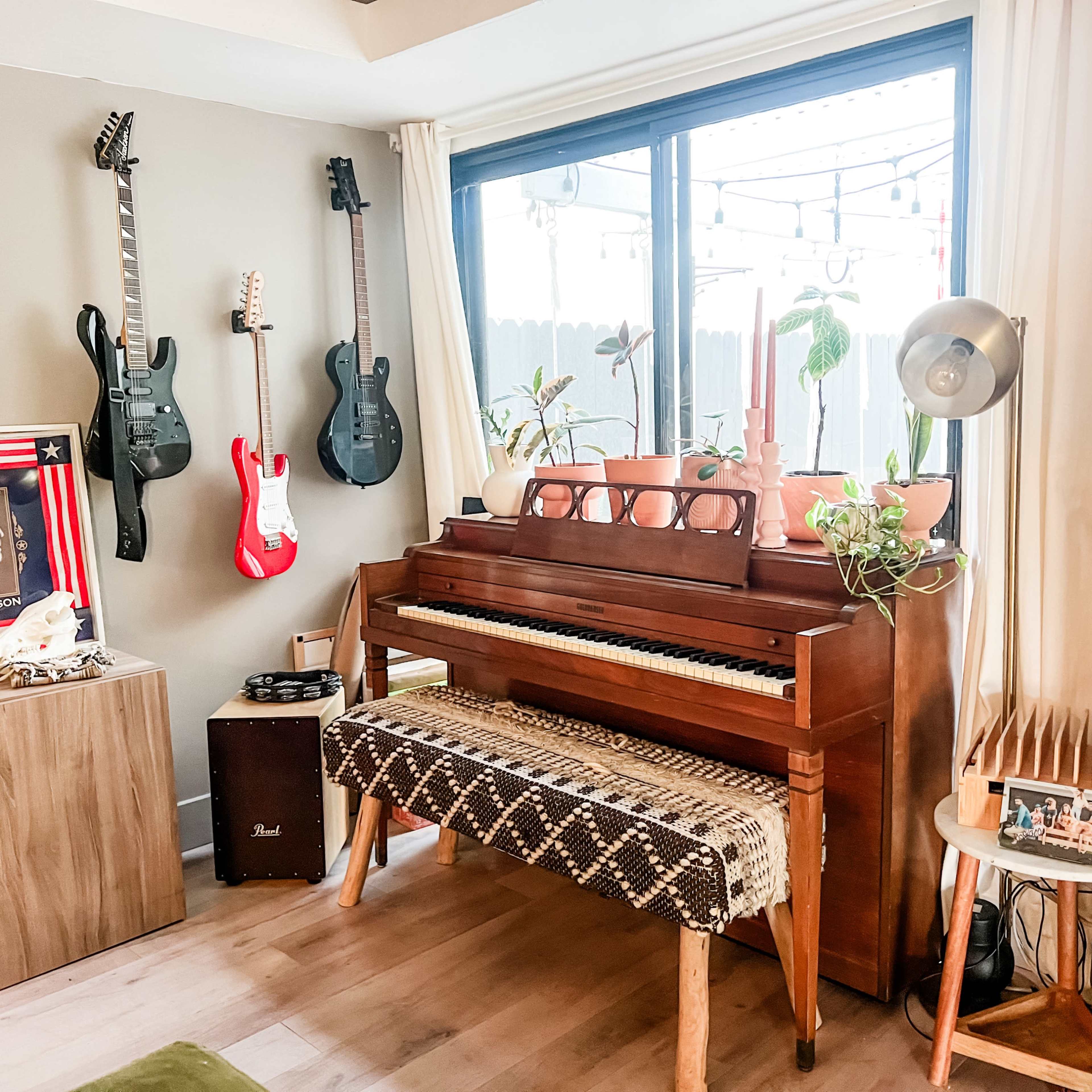 The scene features a wooden piano accompanied by various musical instruments, including guitars and a ukulele, in a well-lit corner with plants and decor.