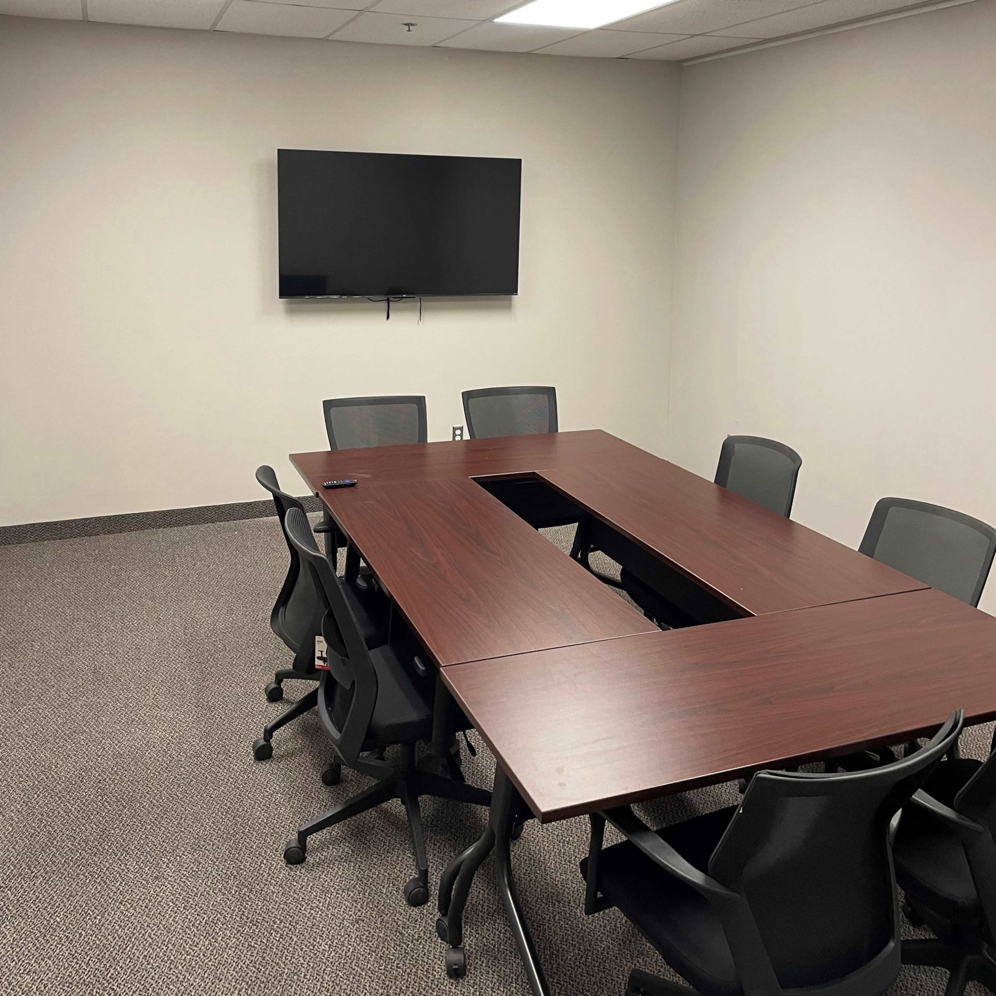 A conference room with a large wooden table surrounded by rolling chairs and a mounted television on the wall.