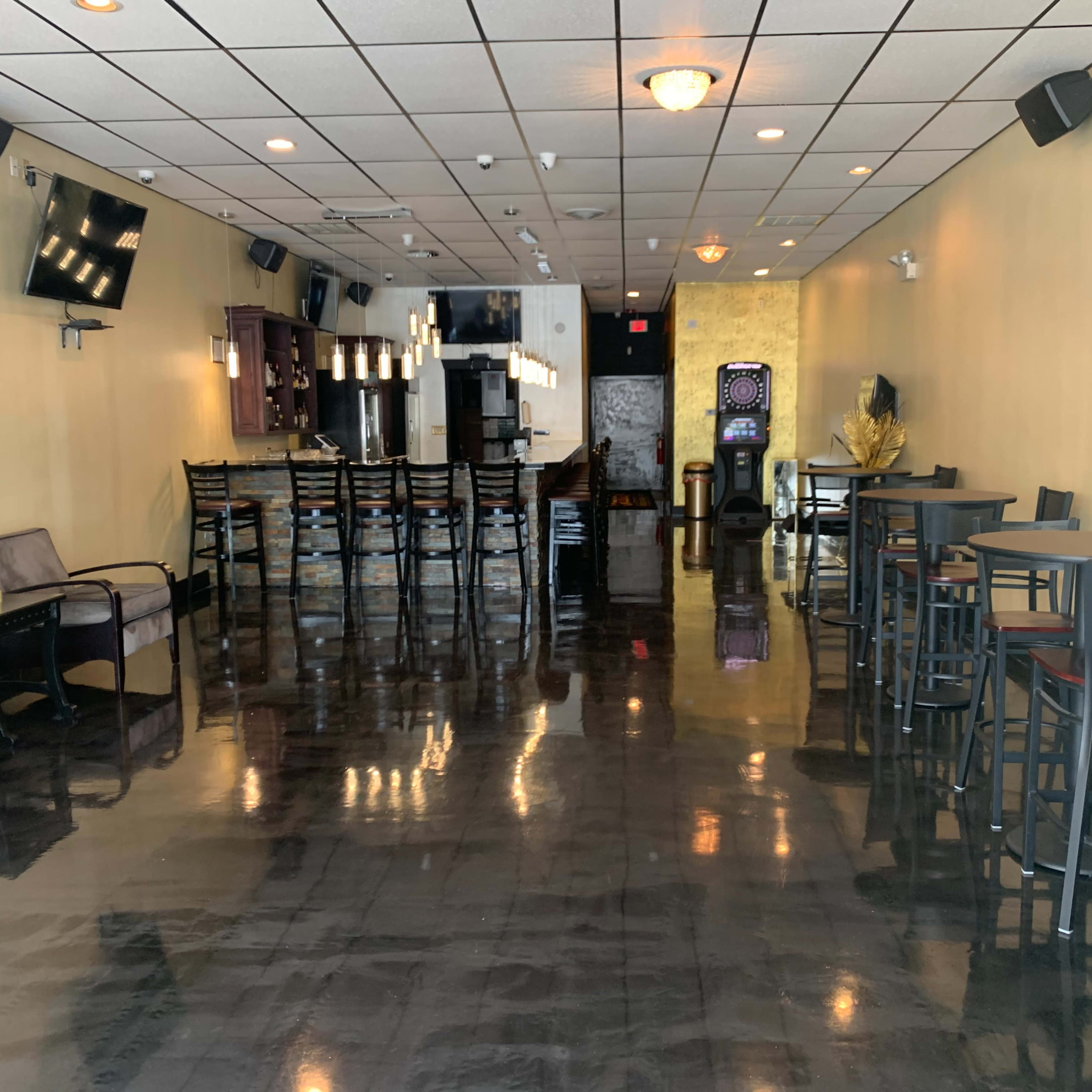 A spacious interior of a modern lounge or bar featuring black polished floors, overhead lighting, a long bar counter, and various seating arrangements.