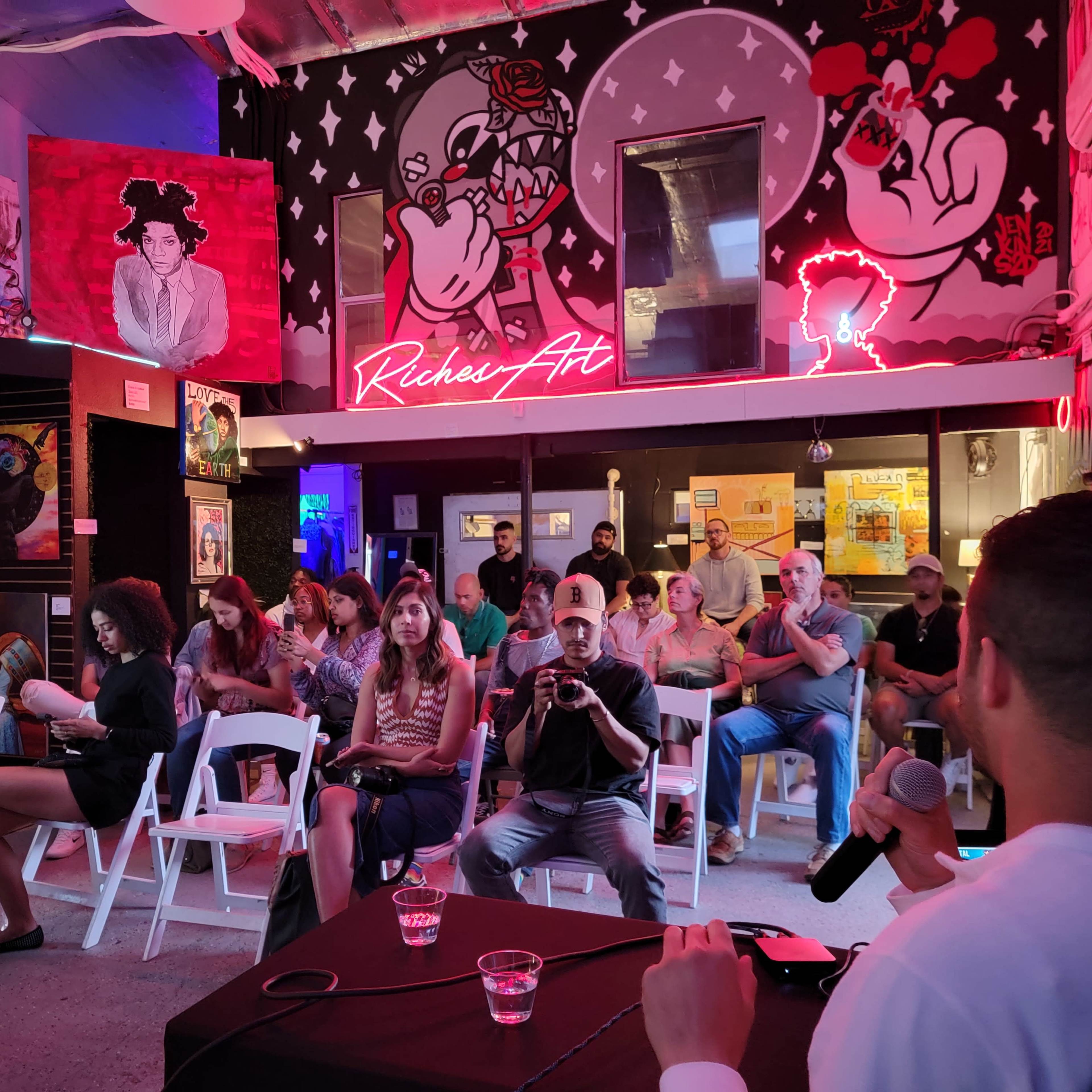 A crowd of people sits in wooden chairs inside an art gallery featuring colorful murals and neon signs, while a speaker addresses them from the front.