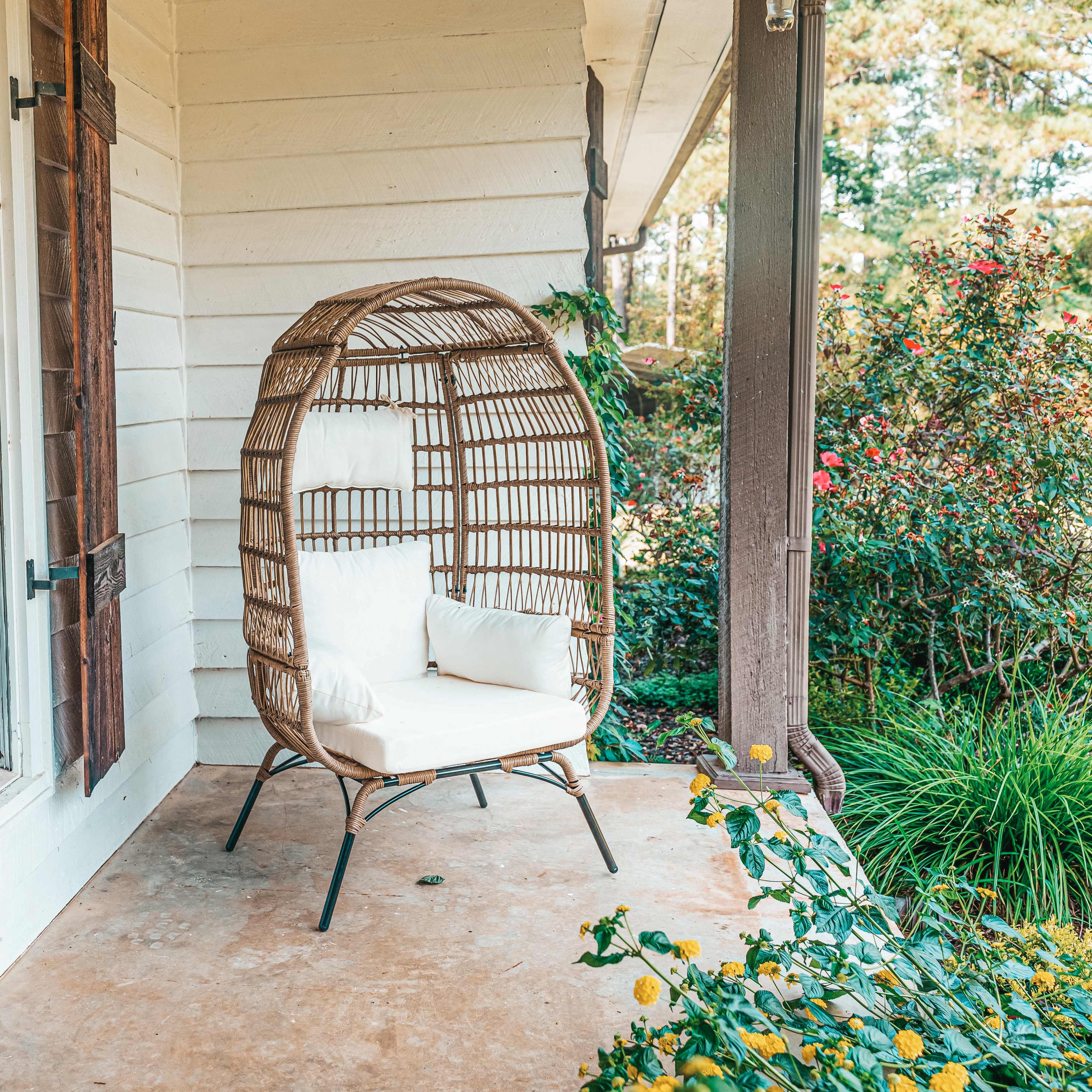 A wicker chair with white cushions is positioned on a porch beside a green shrub and flowers.