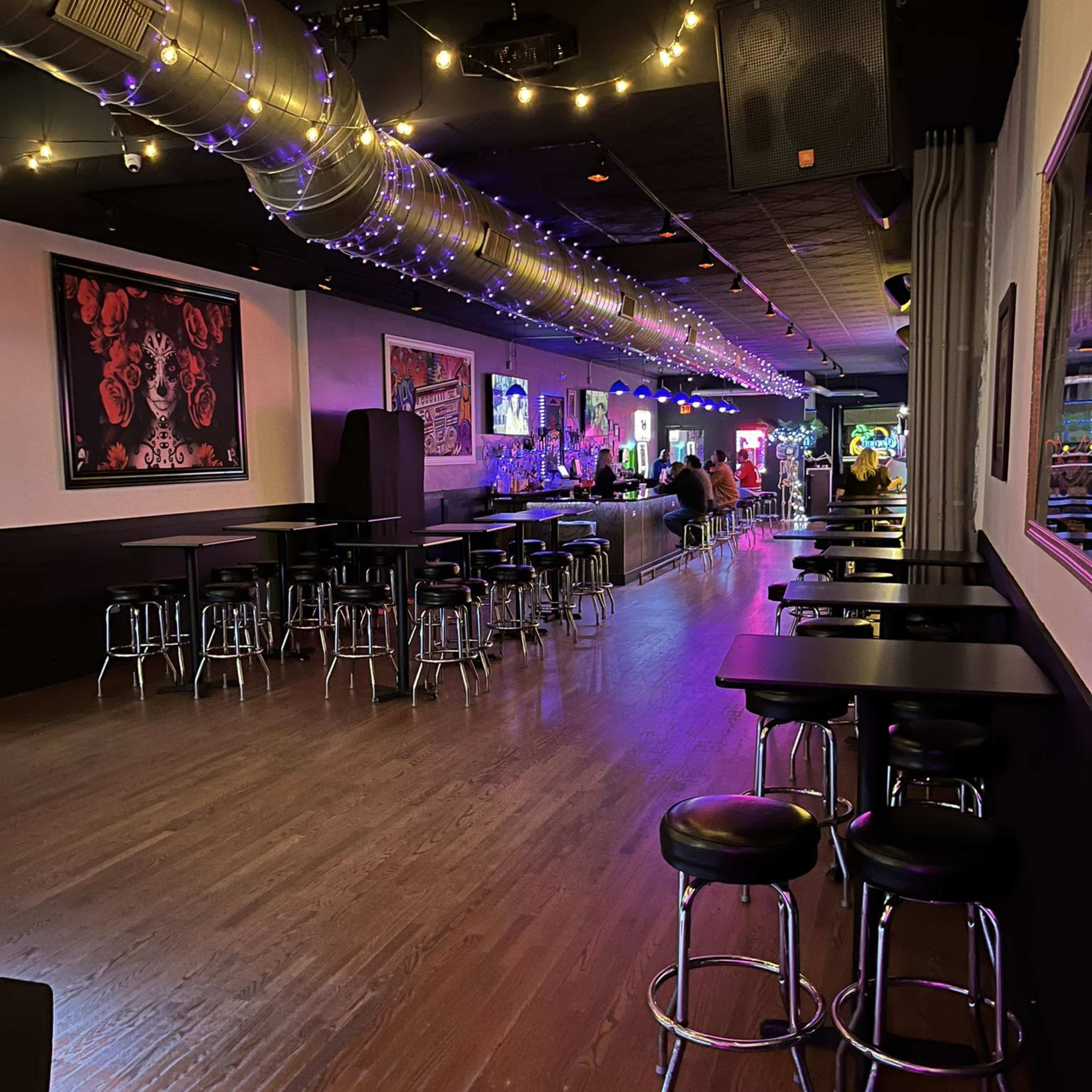 An empty bar area with black tables and stools, illuminated by colorful lights and decorated with artwork on the walls.