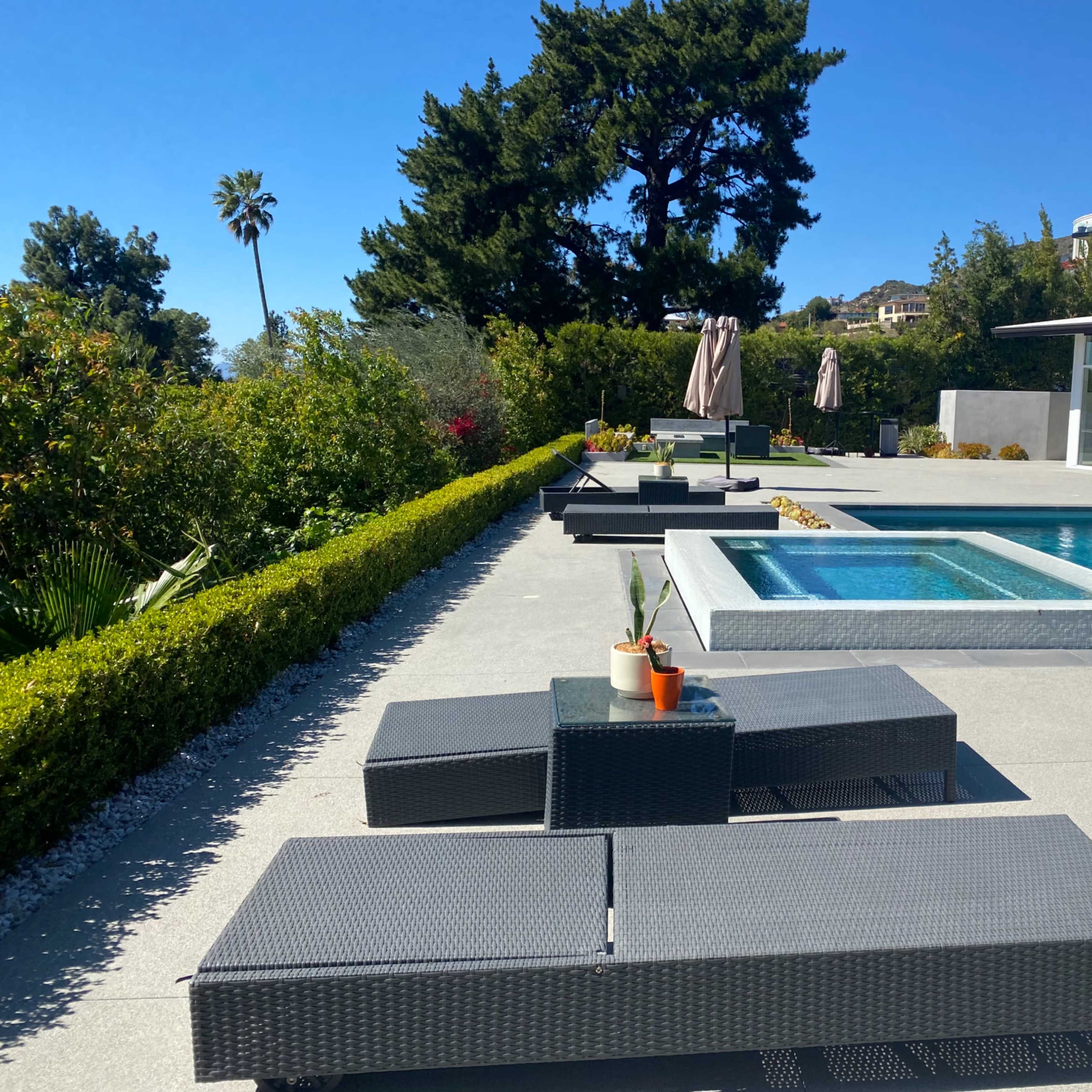 A poolside area features several lounge chairs arranged around a rectangular swimming pool with a spa, set against a backdrop of lush greenery and palm trees.