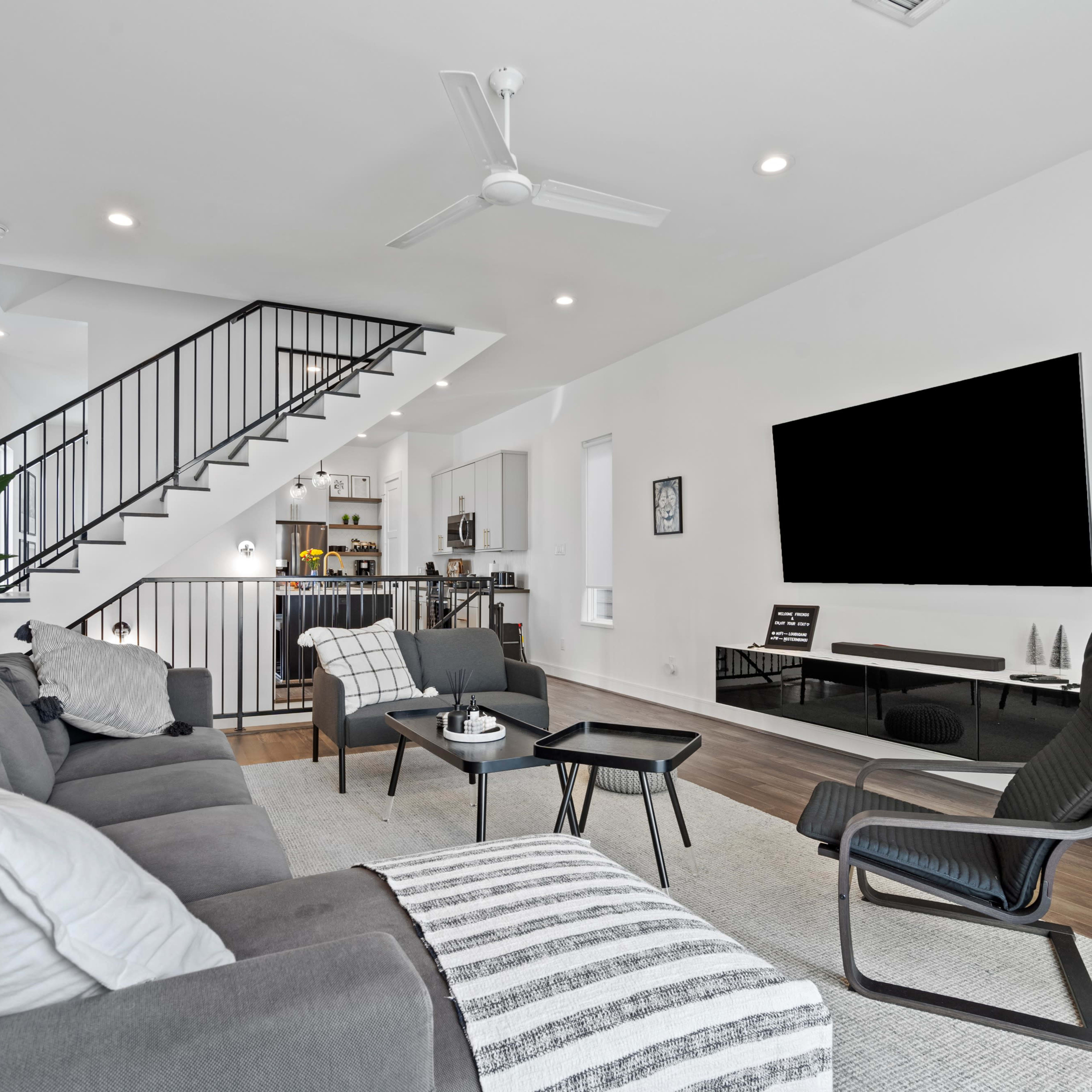 A modern living room features a gray sectional sofa, a striped area rug, and a large wall-mounted television, with a staircase visible in the background leading to a kitchen area.