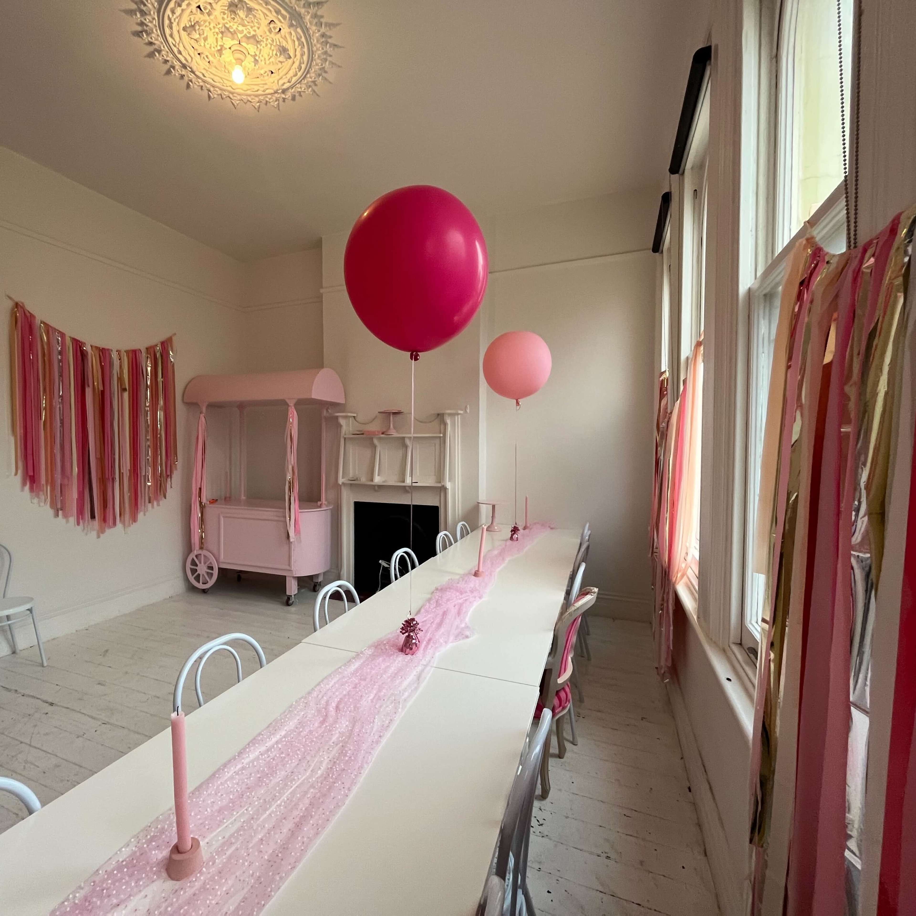 A long table is set with a pink tablecloth and candles, flanked by colorful streamers and two large pink balloons in a bright room.