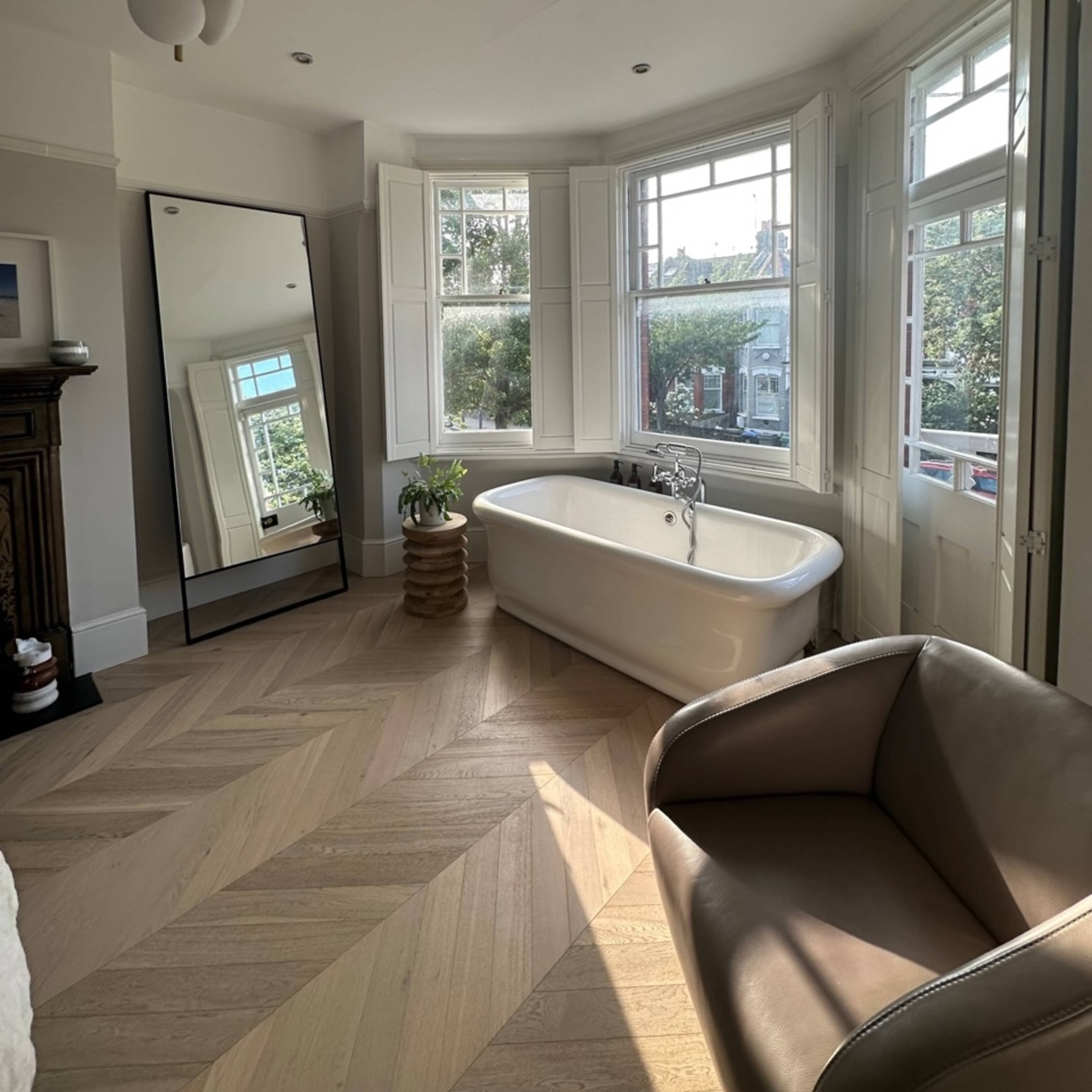 A bright bathroom features a freestanding bathtub, a large mirror, and bay windows overlooking a green space.