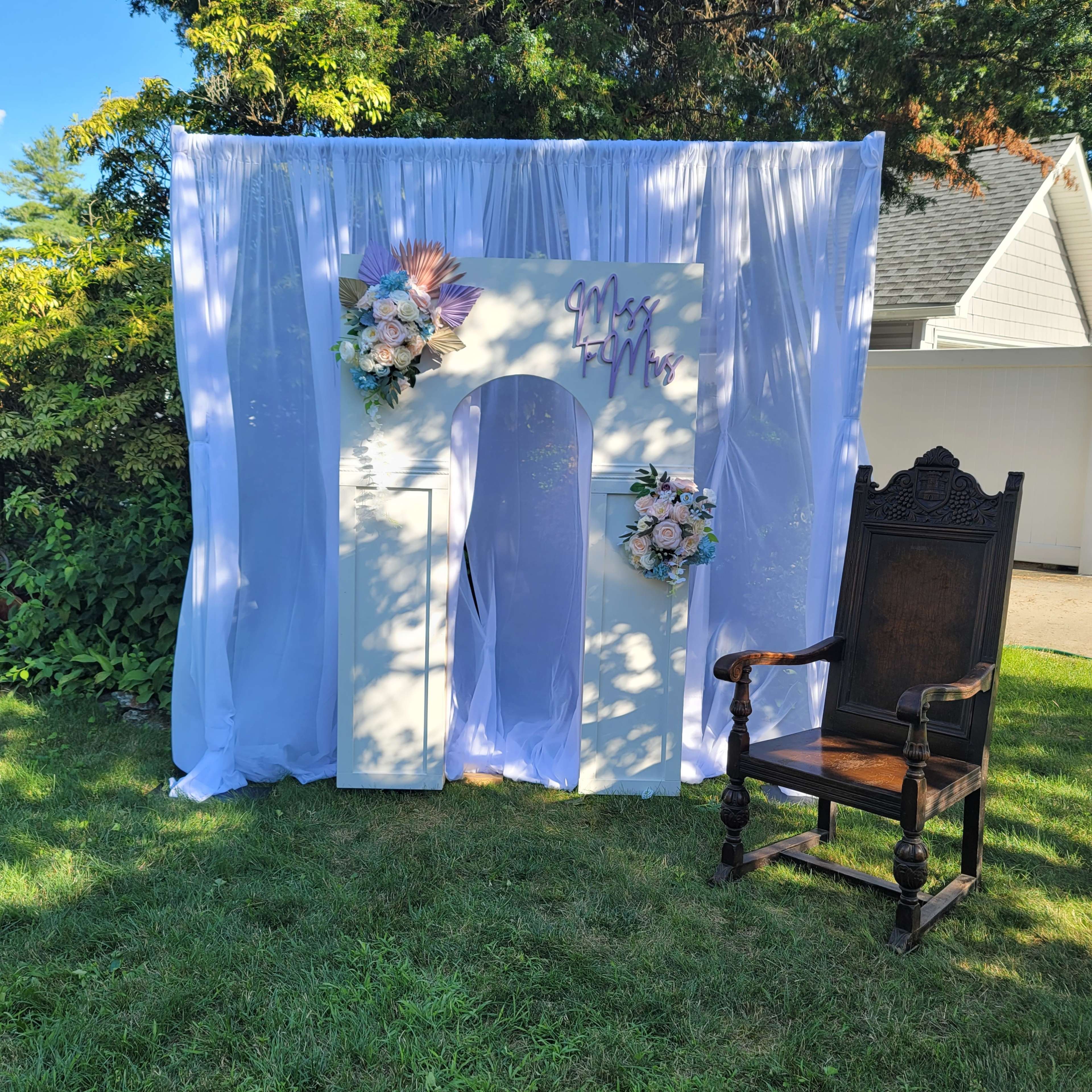 A decorated backdrop with white drapes, floral arrangements, and a sign reading "Miss FUN," accompanied by an ornate wooden chair on a grassy area.