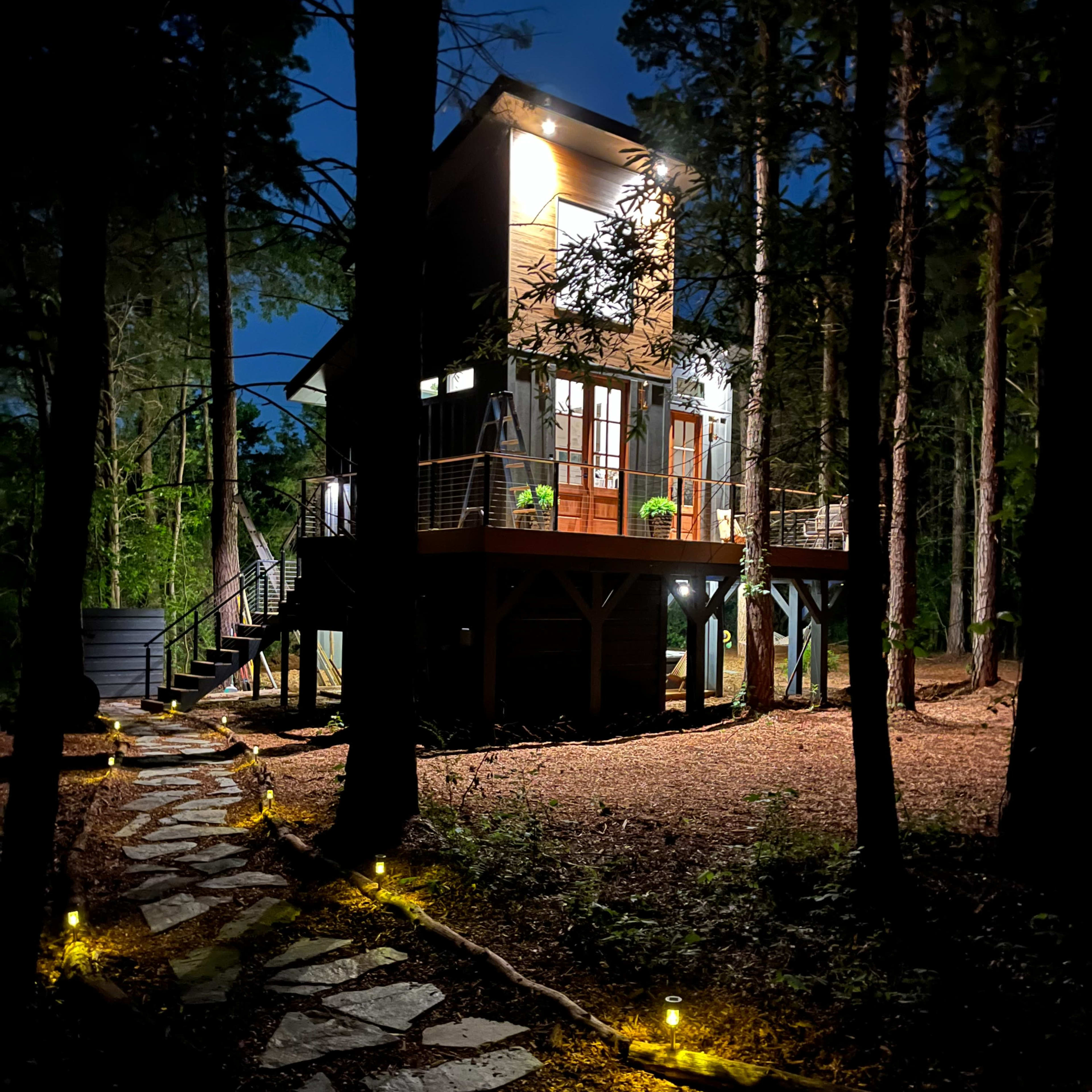 A modern two-story cabin is illuminated at night, surrounded by tall trees and a stone pathway lined with solar lights.