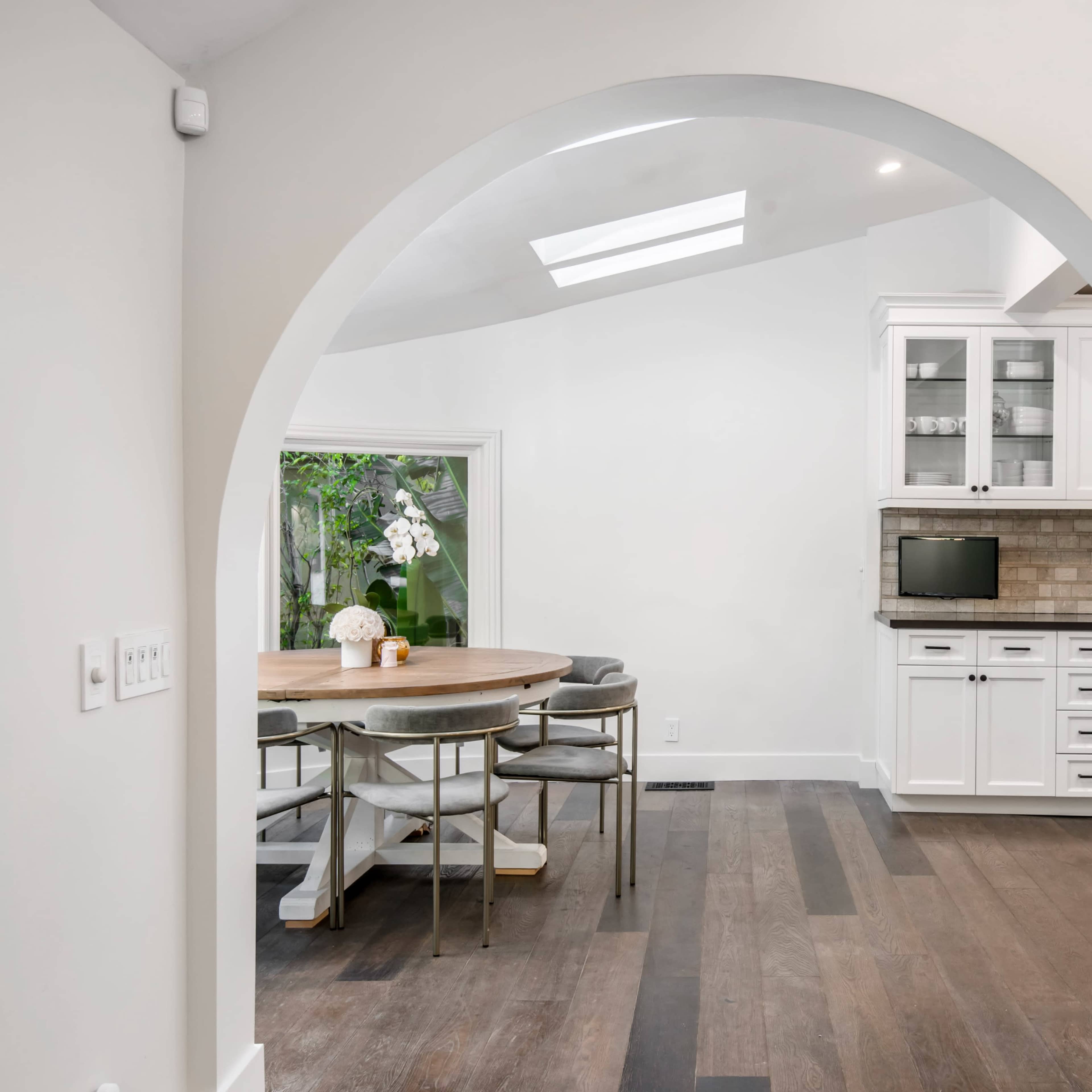 A modern kitchen with white cabinetry and a round wooden dining table is visible through an arched passageway.