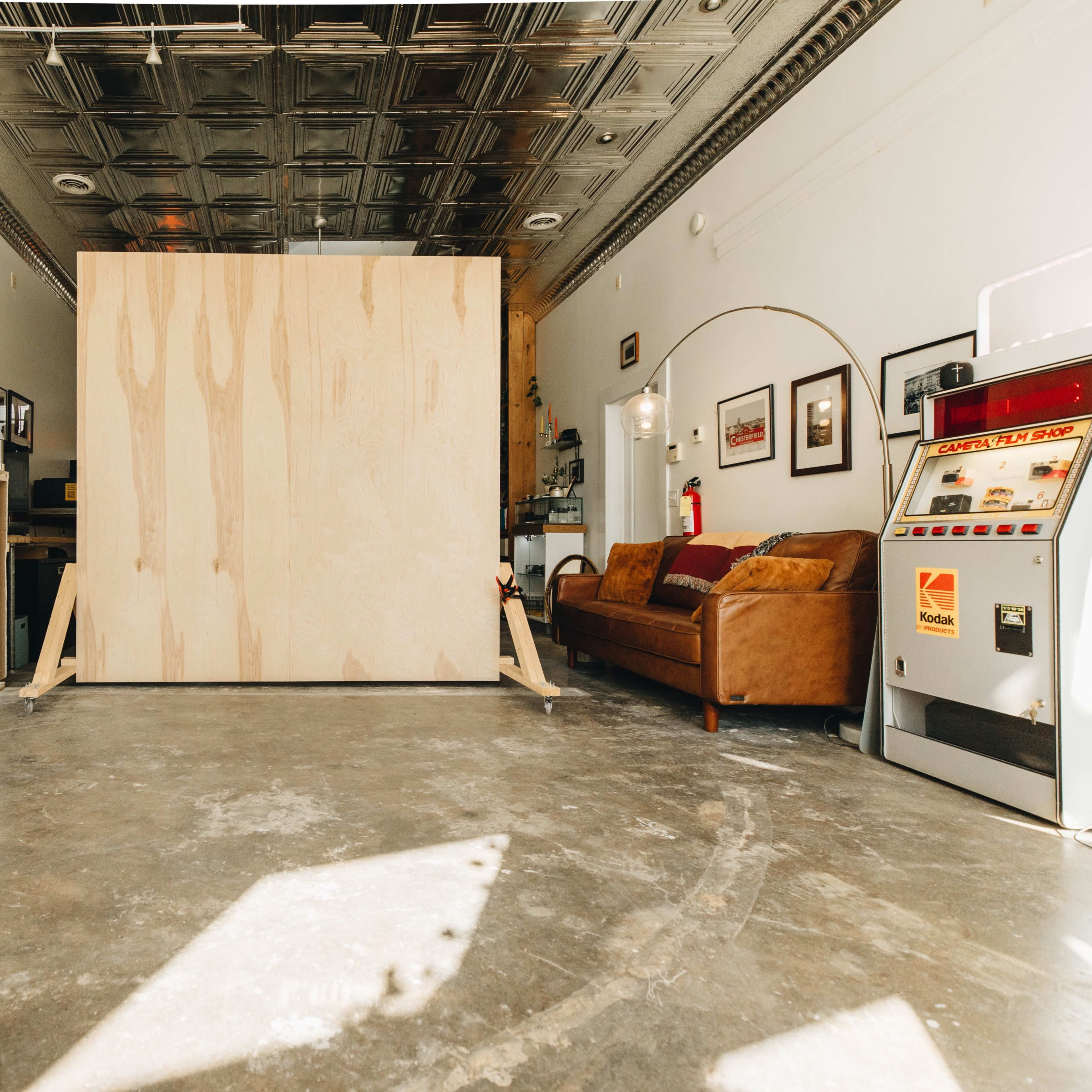 A large plywood panel stands in the center of a spacious room with a concrete floor, flanked by a mirror, a couch, and various items against the walls.