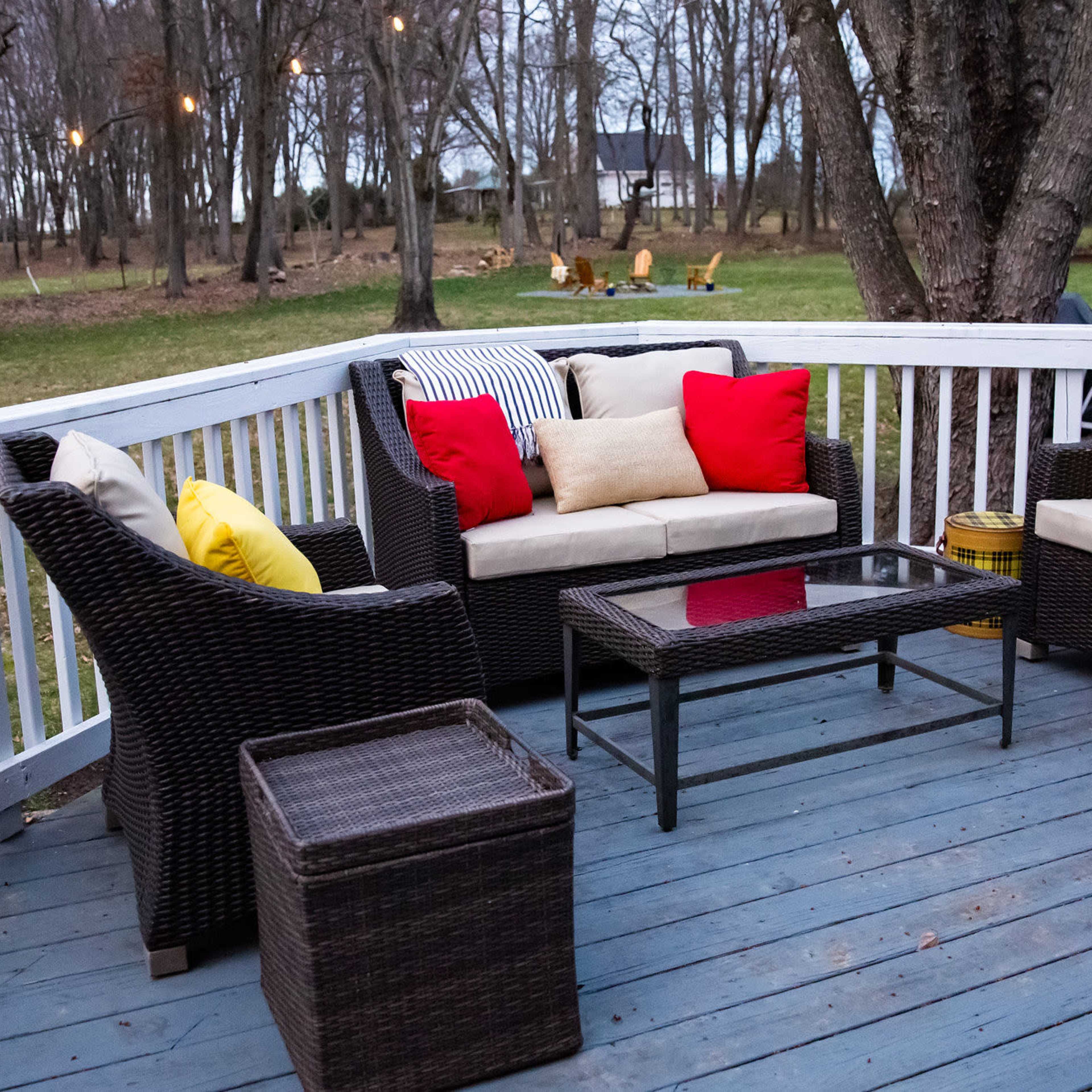 A patio deck features a seating area with wicker furniture, including a coffee table, colorful cushions, and a small side table, surrounded by trees.
