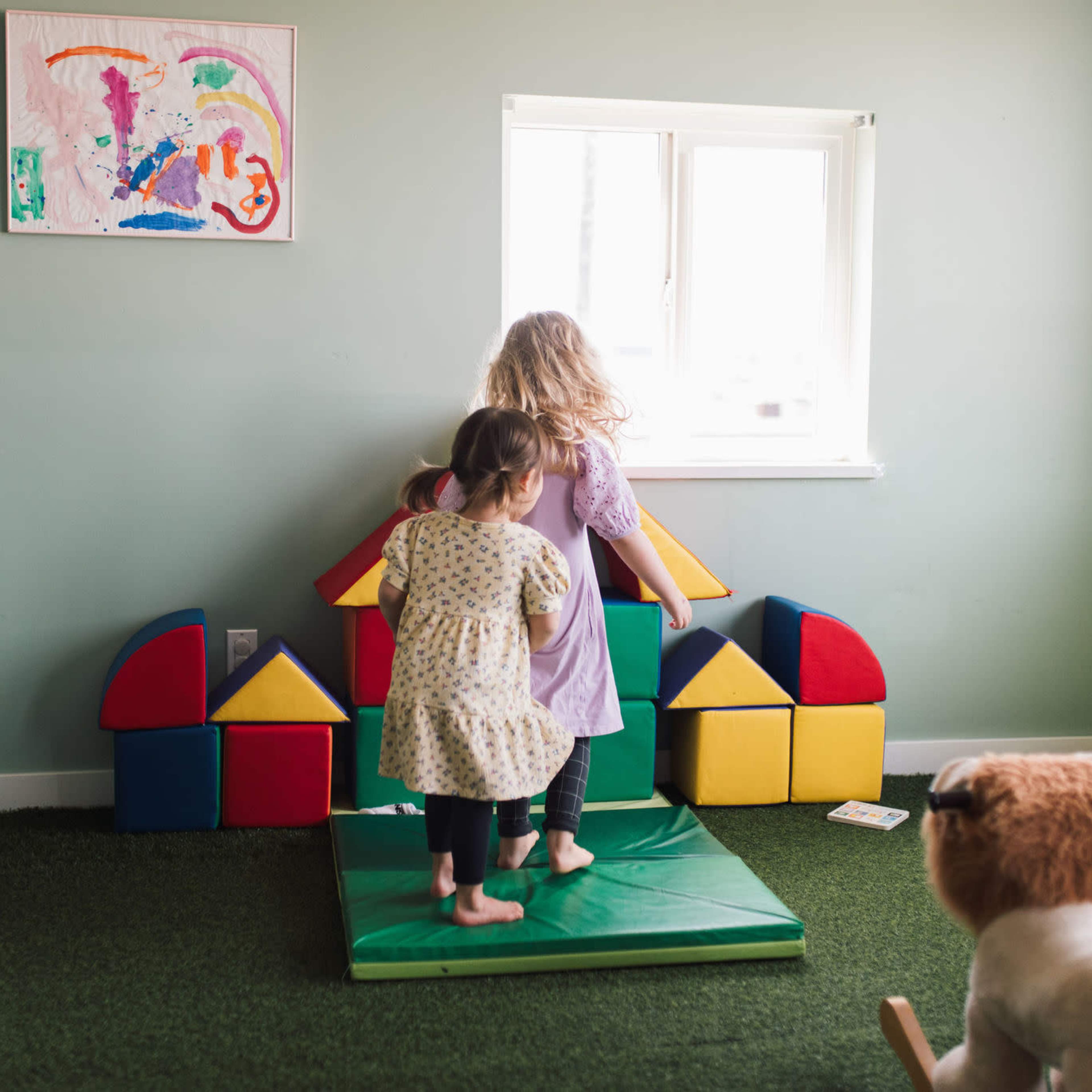 Two children stand on a green mat in a brightly colored playroom with building blocks arranged in the shape of a house and a window in the background.