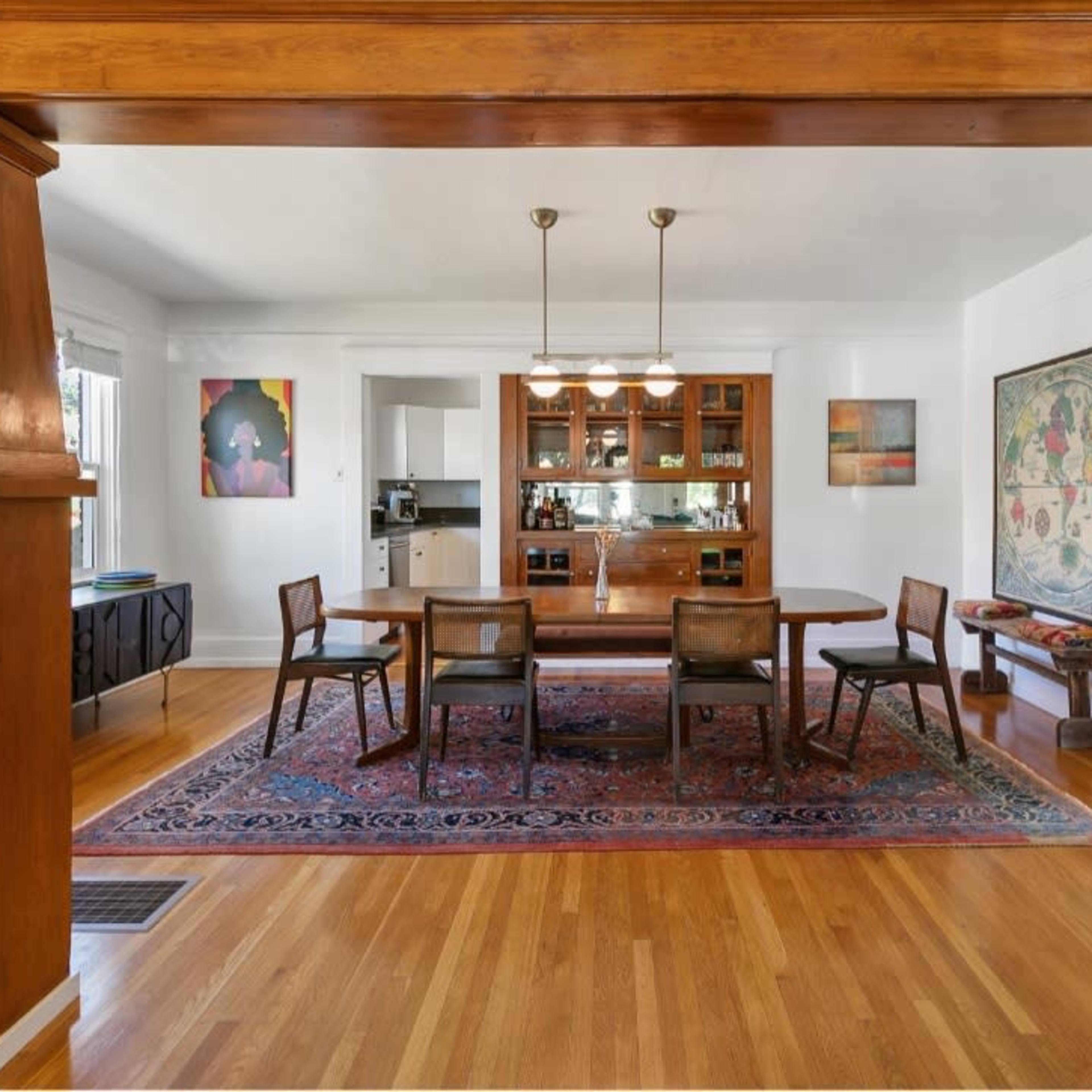 A dining area features a wooden table surrounded by chairs, with a sideboard and a kitchen visible in the background.