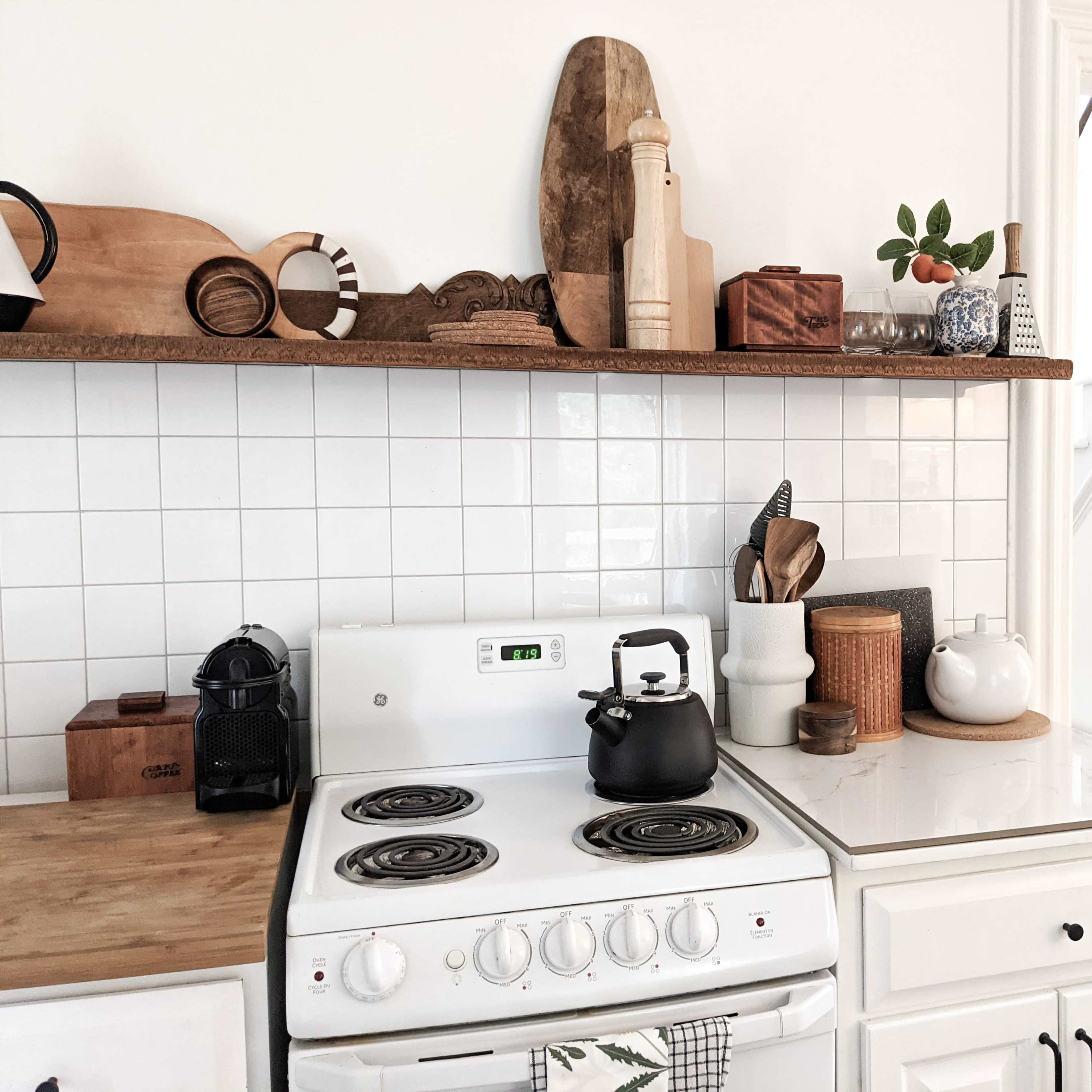 A modern kitchen features a white gas stove, wooden shelves displaying decorative items, and a mix of cooking utensils and containers on the countertop.