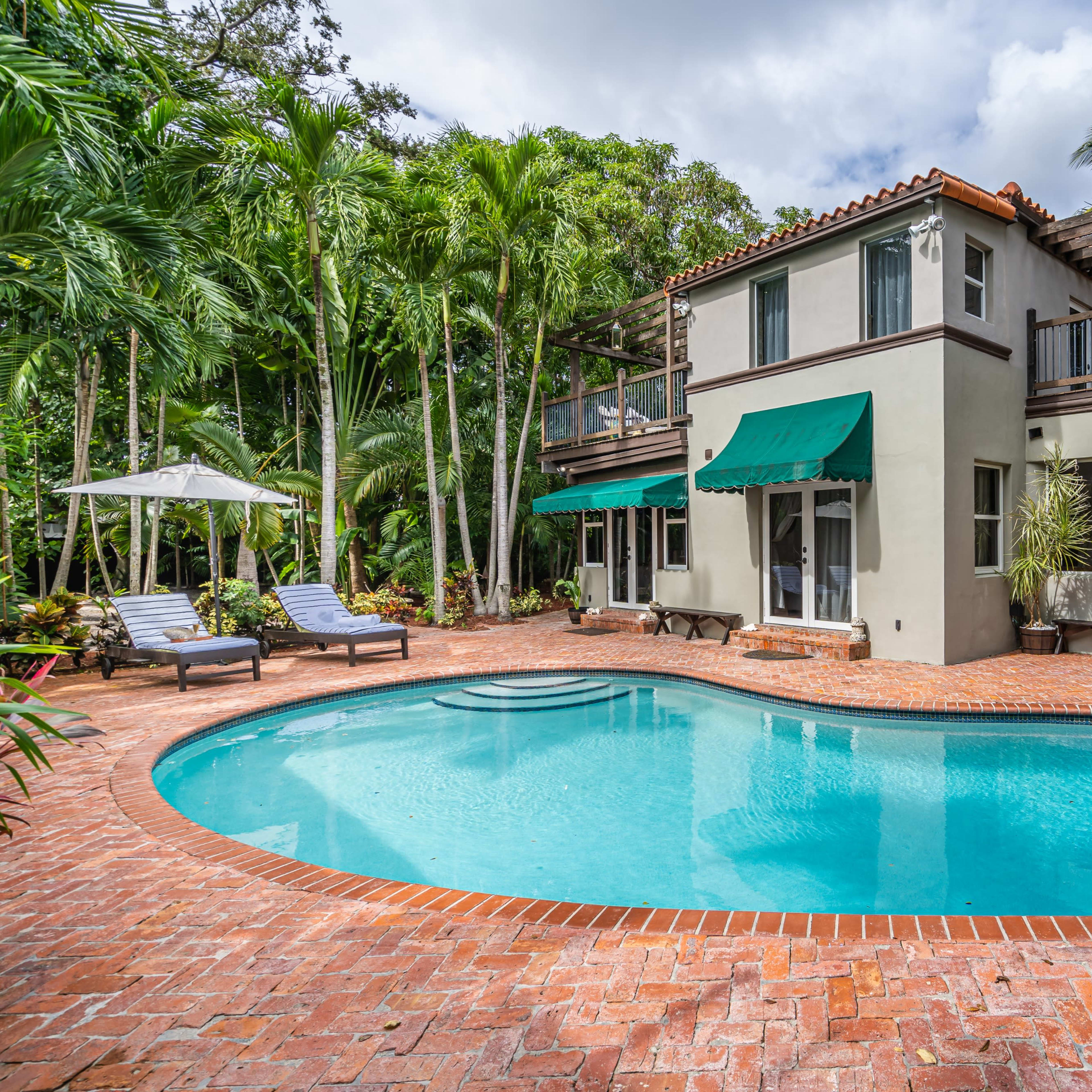 A two-story house with green awnings, surrounded by palm trees, featuring a swimming pool with a brick patio.