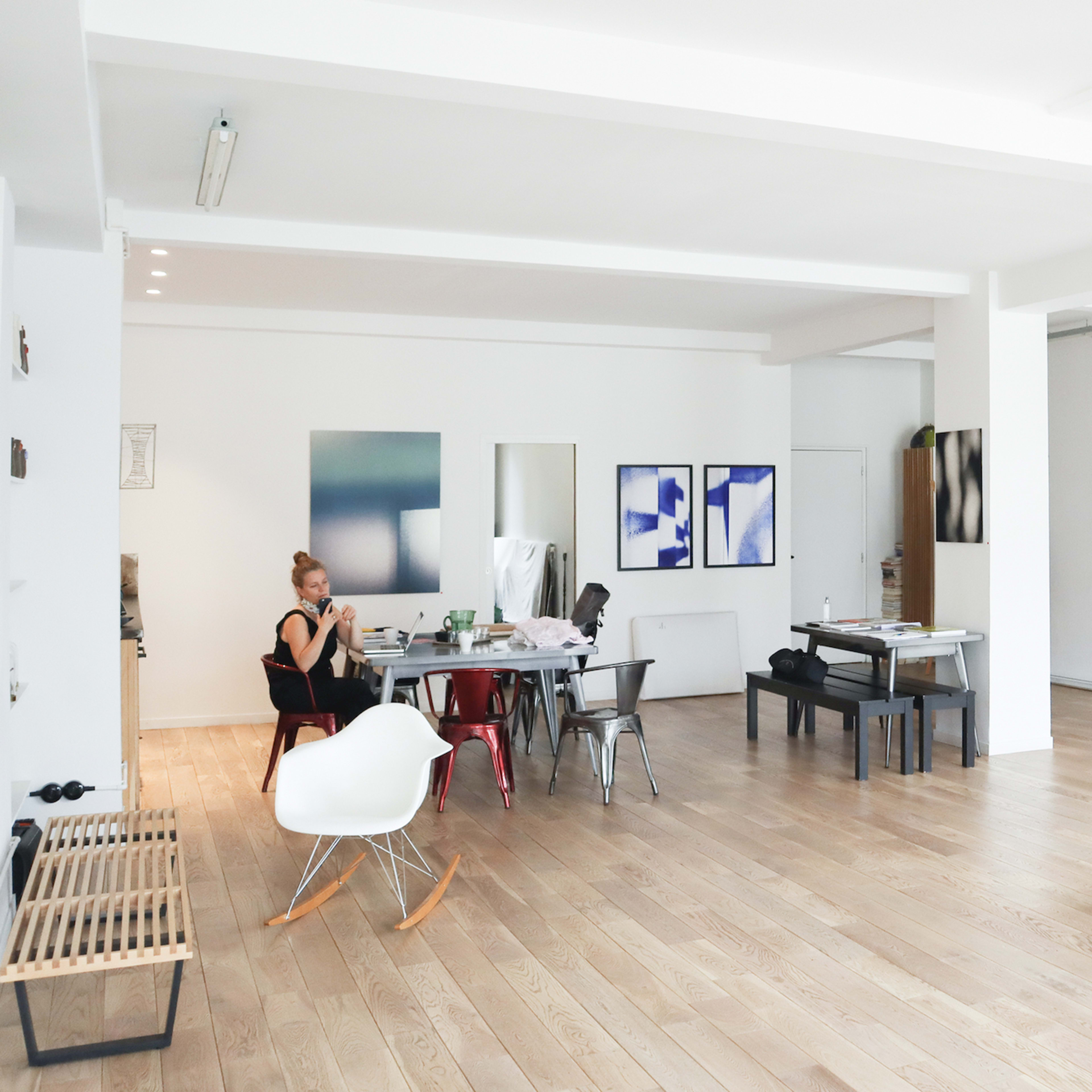 A spacious, modern art studio with a woman sitting at a table, surrounded by various artworks and furniture.