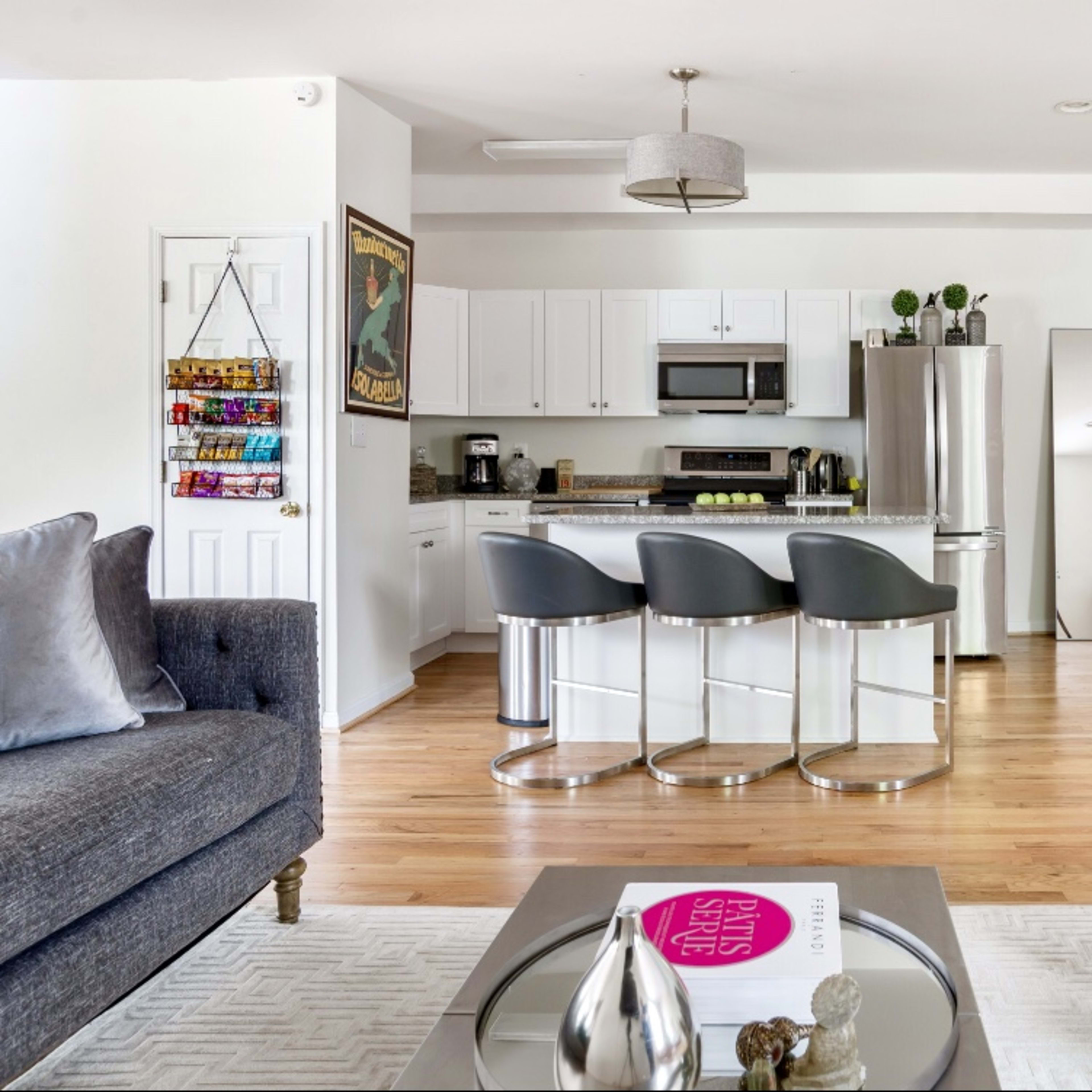 A modern living room with a gray sofa and armchair, a coffee table with a magazine, and a kitchen area featuring white cabinets and bar stools.
