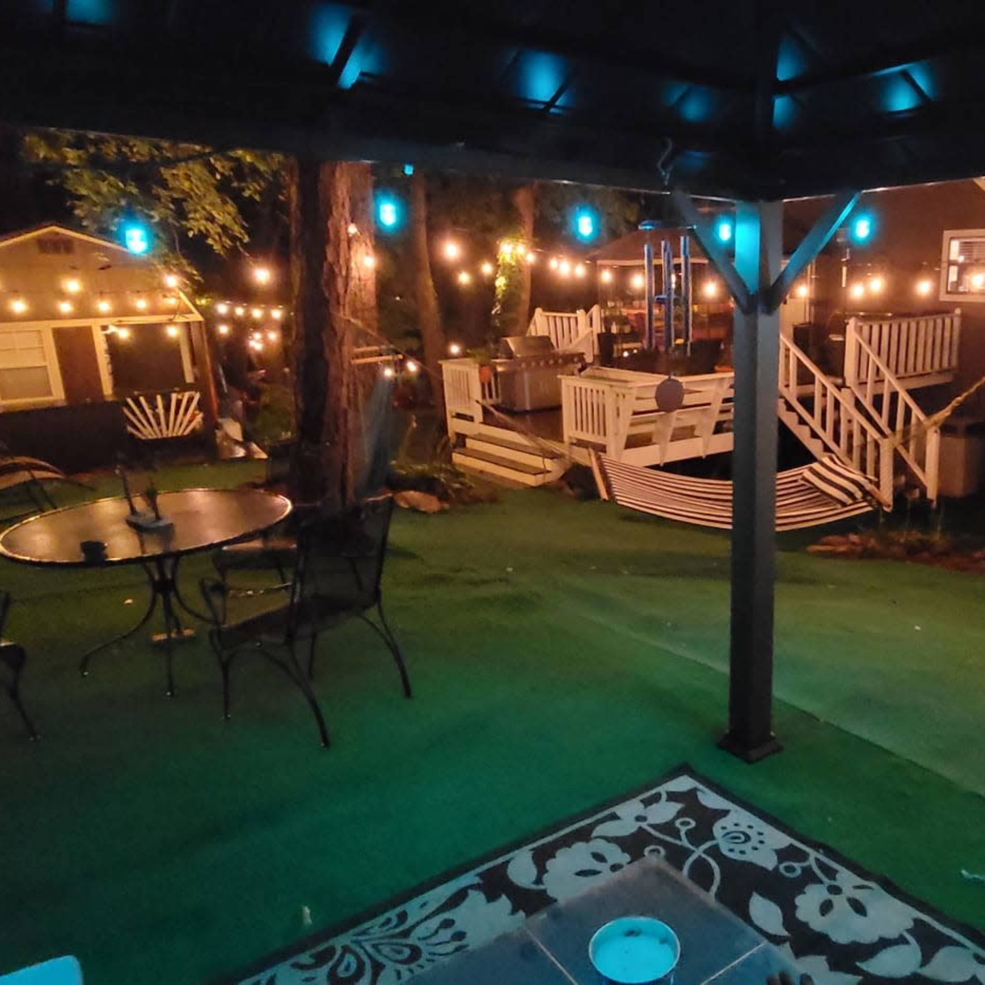 The scene shows a backyard decorated with string lights, featuring a patio area with a table and chairs, along with several small buildings visible in the background.