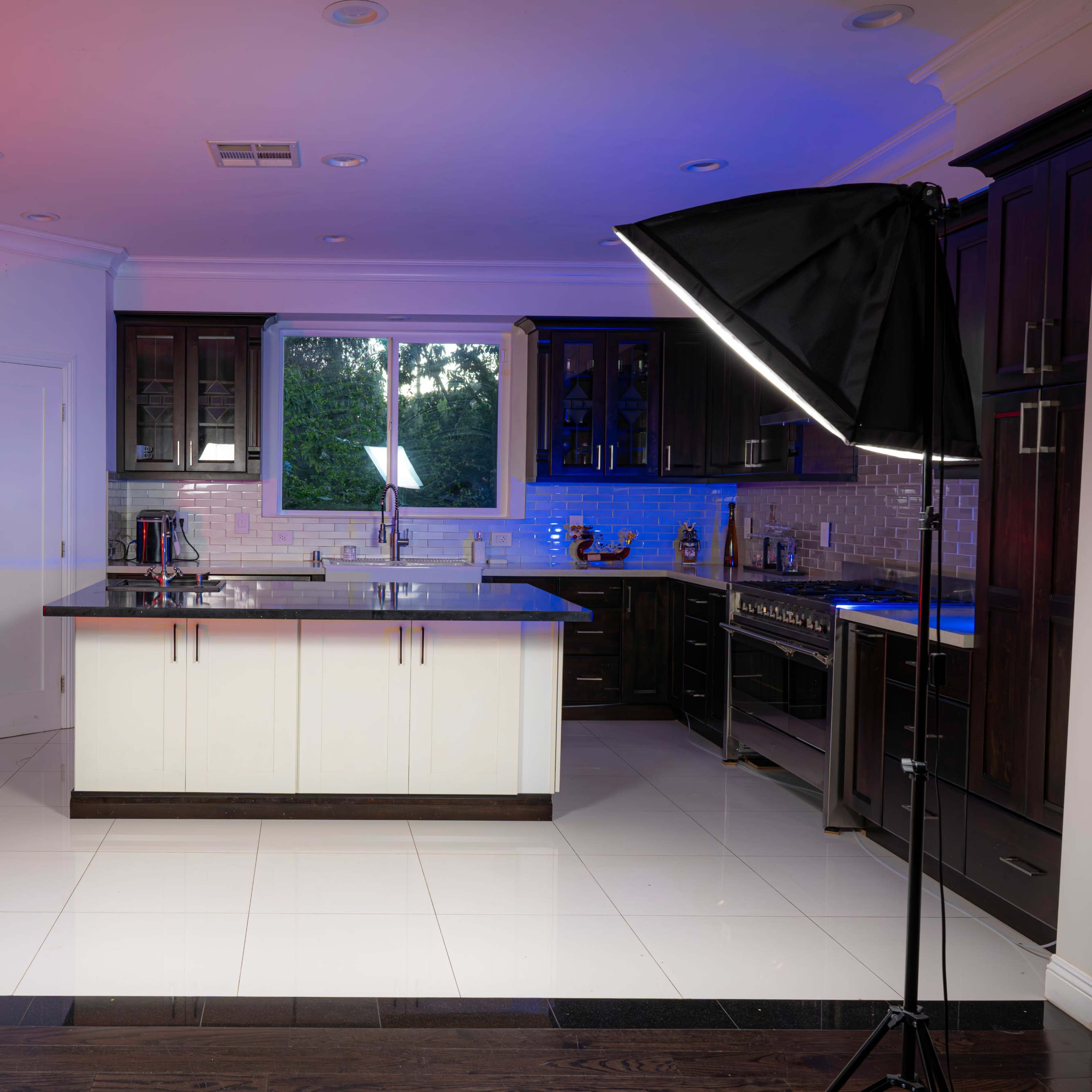 A modern kitchen is illuminated by soft box lights, featuring dark cabinetry, a center island, and stainless steel appliances.