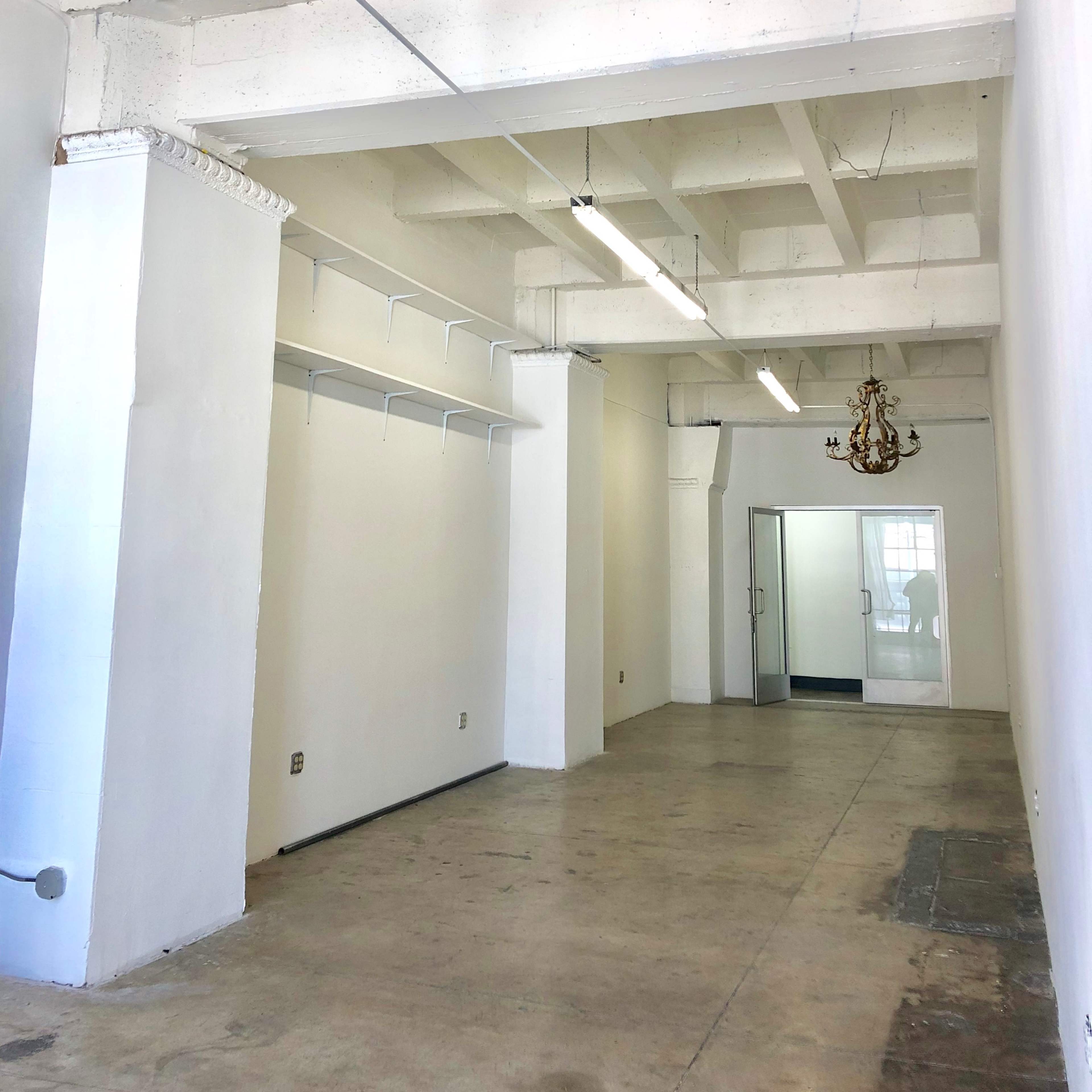 A spacious, empty room with concrete flooring, white walls, and exposed ceiling beams, featuring a chandelier and a glass door leading to another area.